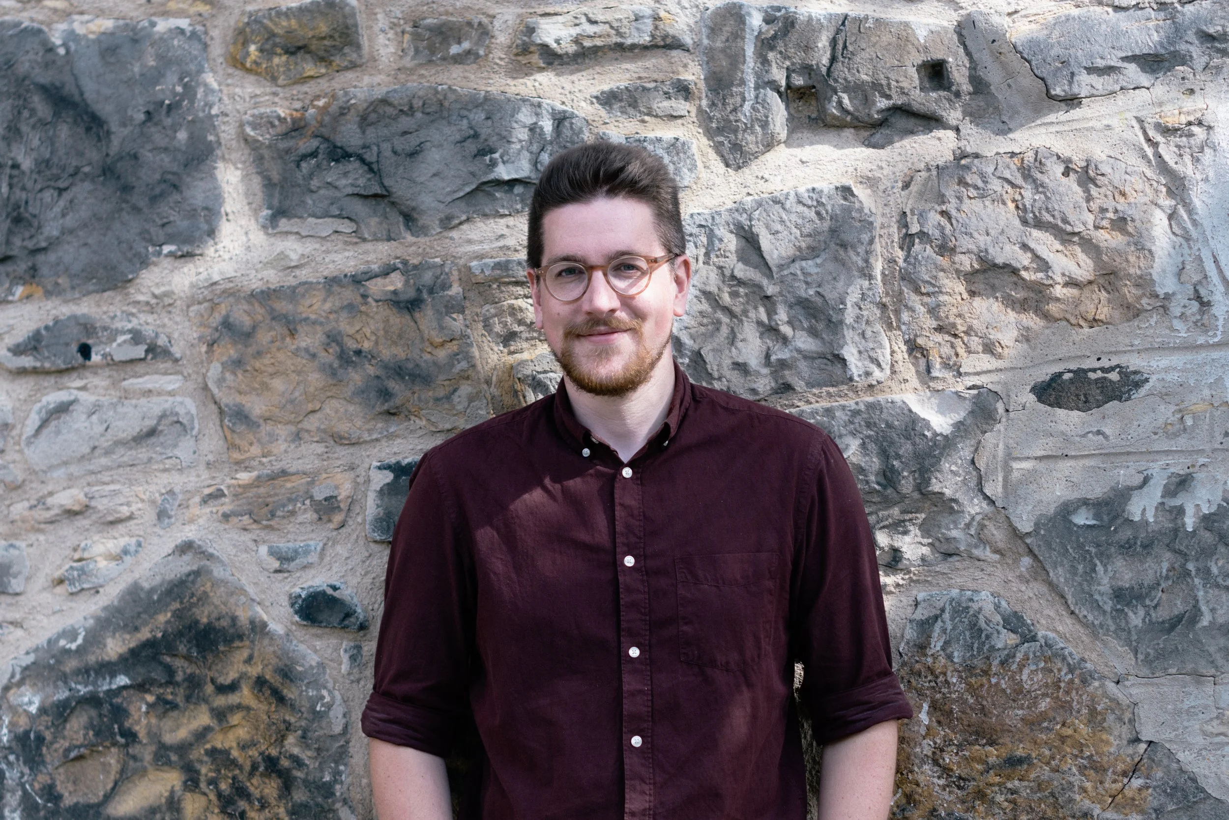 A young man with glasses, a beard, and brown hair smiling in front of a stone wall.