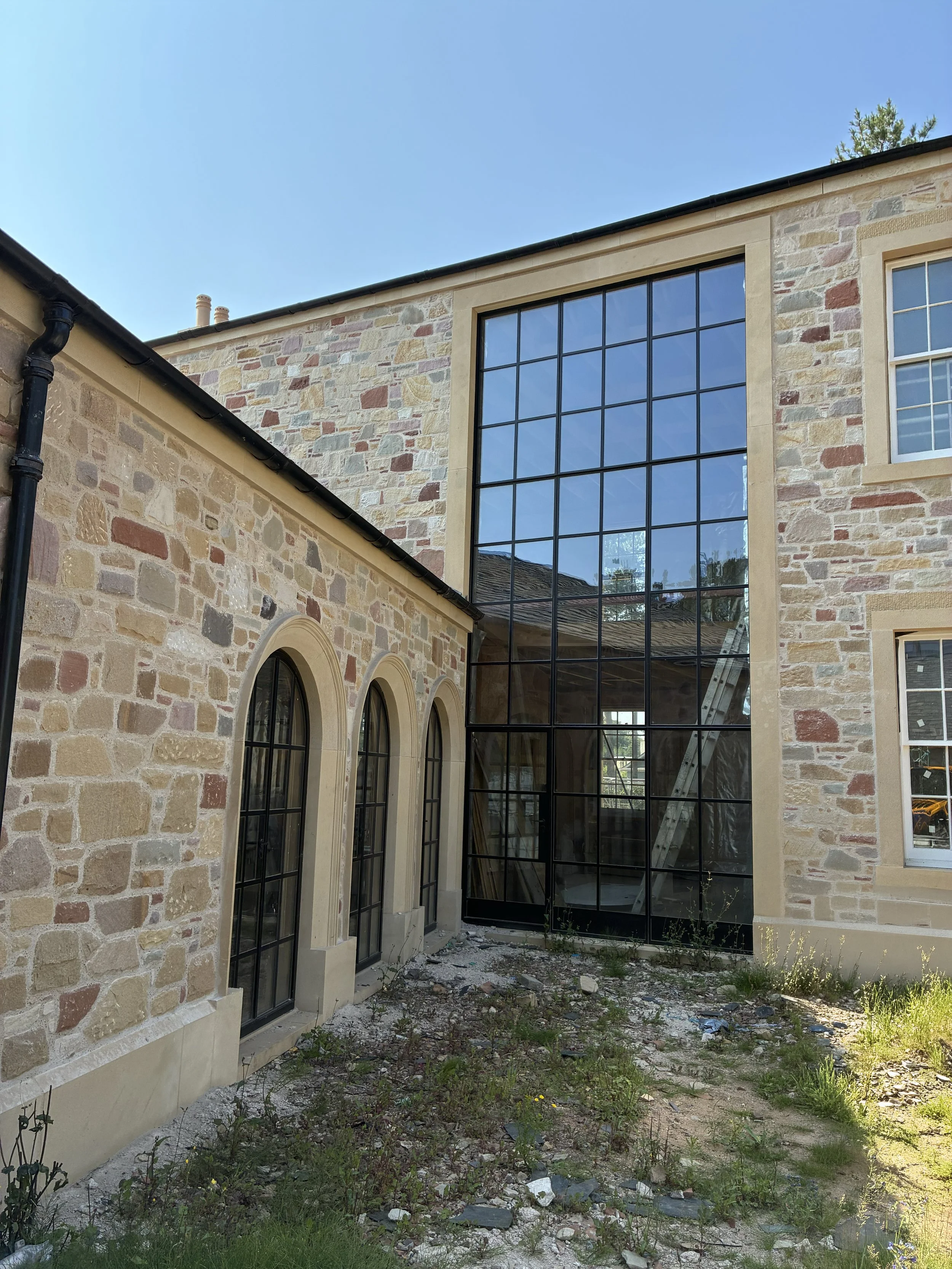A building with a stone exterior and large black-framed glass windows, with an outdoor area that has dirt and sparse grass, and a ladder leaning against the building under a clear blue sky.