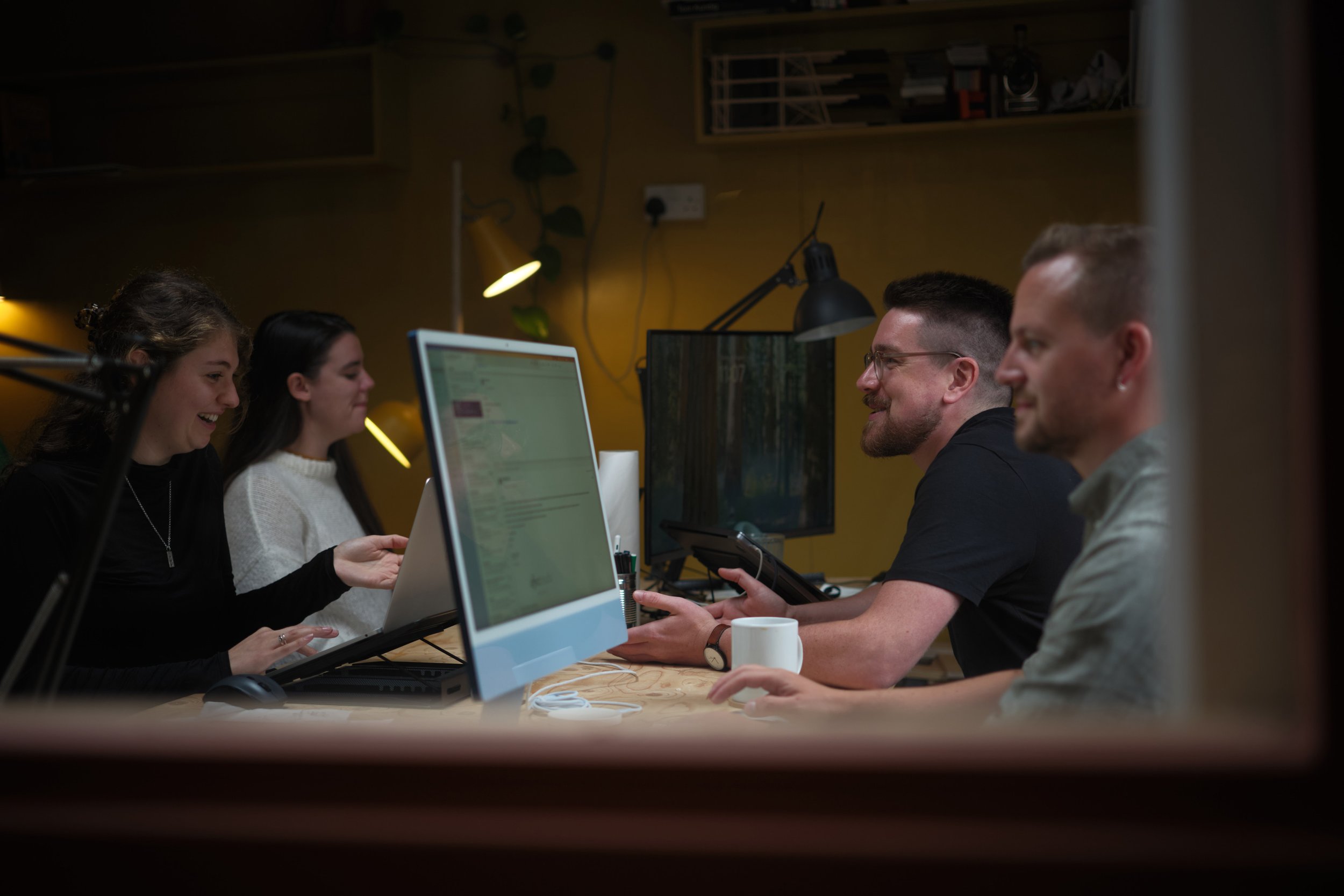 People sitting around a table working on computers in a modern office, visible through a narrow framed opening.