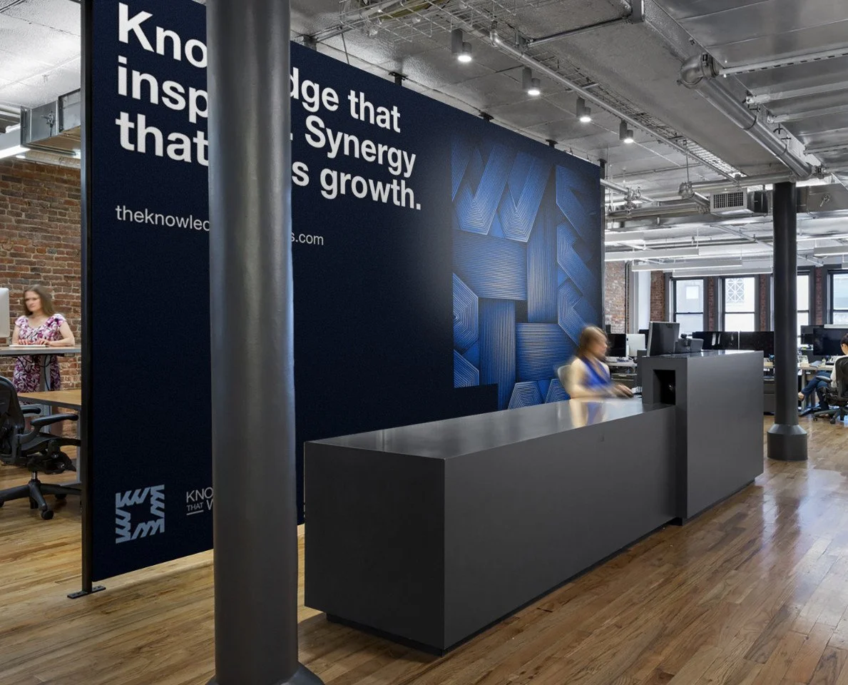 Modern office reception area with a dark gray desk, large wall mural displaying the text 'Knowledge that inspires. Synergy that drives growth' and 'theknowledges.com,' open workspace with exposed brick and industrial decor.