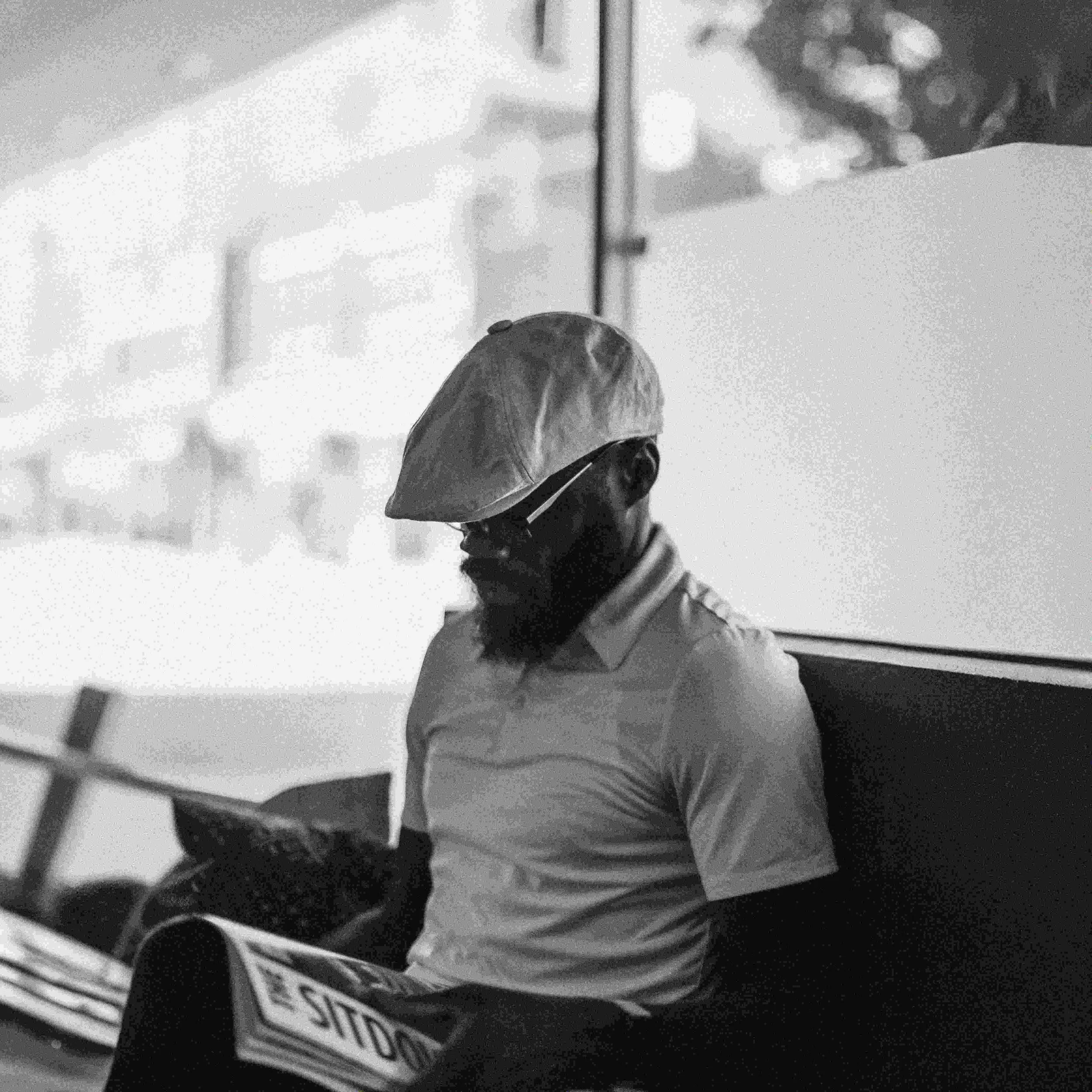 Black and white photo of a man with a beard, wearing a hat and sunglasses, sitting and reading a newspaper.