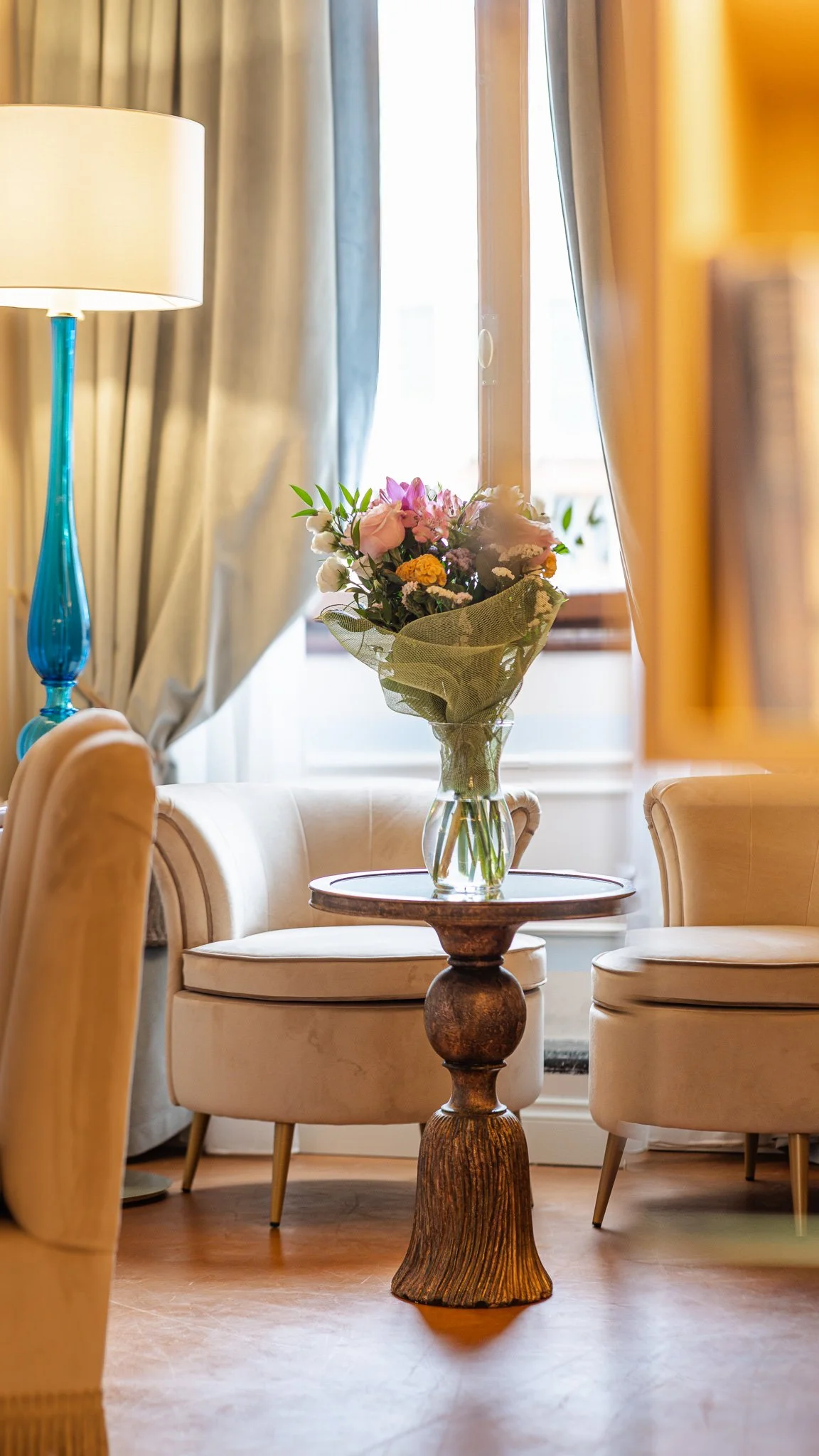 Vaso di fiori freschi su un tavolino in una stanza decorata con tende color crema e una lampada blu.