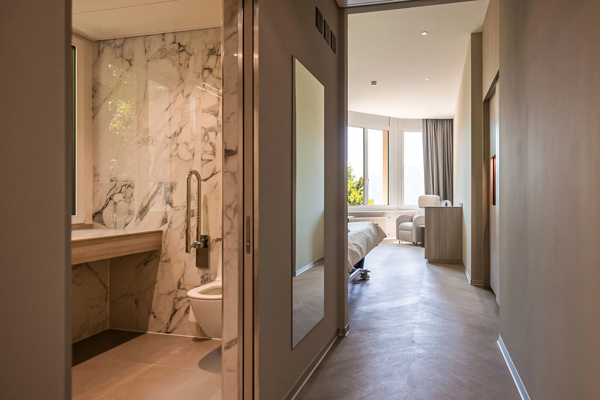 Vista di un corridoio di un hotel con una camera da letto visibile in fondo e un bagno a sinistra, con pareti chiare e grandi finestroni che lasciano entrare molta luce naturale.