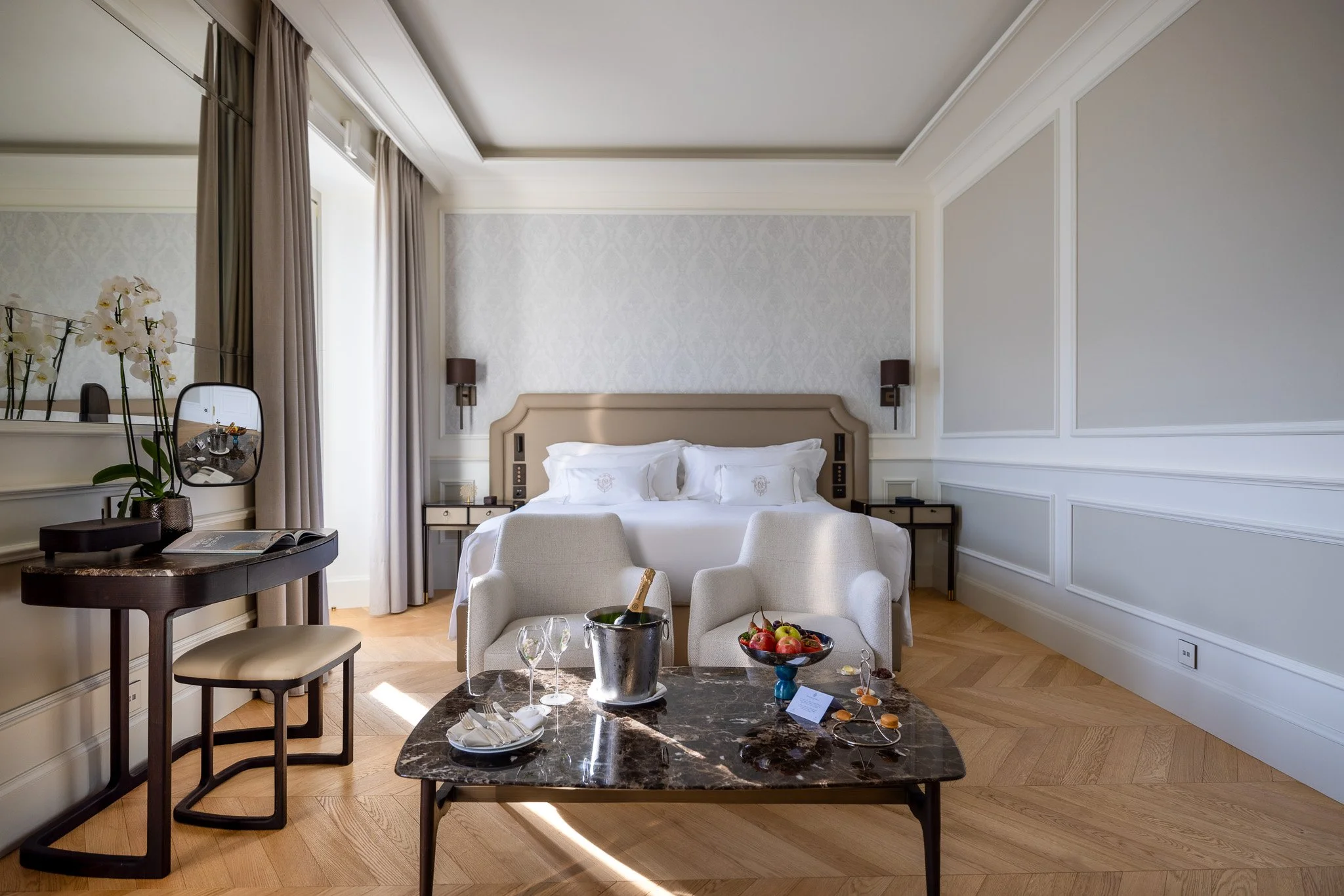 Camera d'hotel elegante con letto matrimoniale, poltrone, tavolo con bicchiere di champagne e frutta.