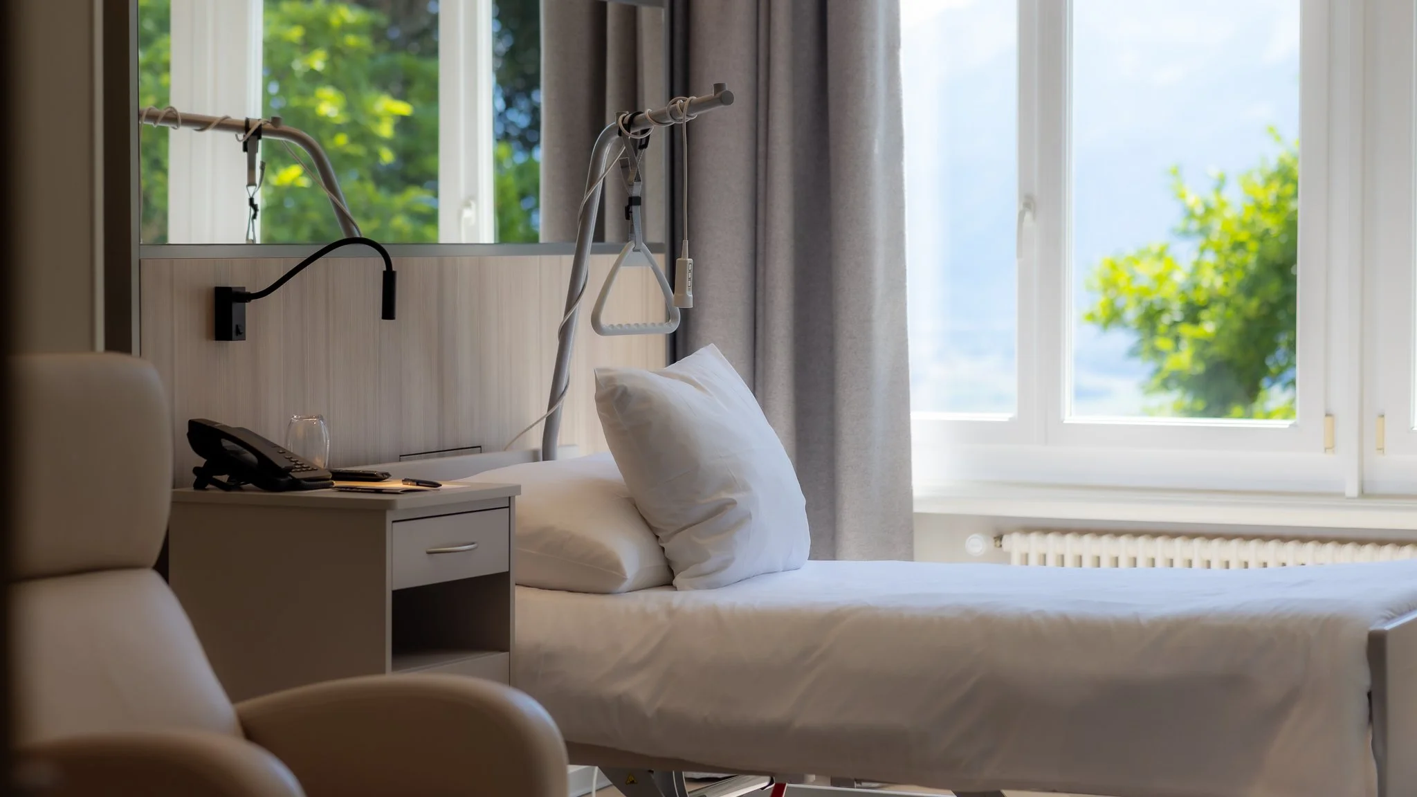 Letto in una stanza d'ospedale con finestra aperta che mostra alberi verdi e cielo blu, presenza di telefono sulla comodina.