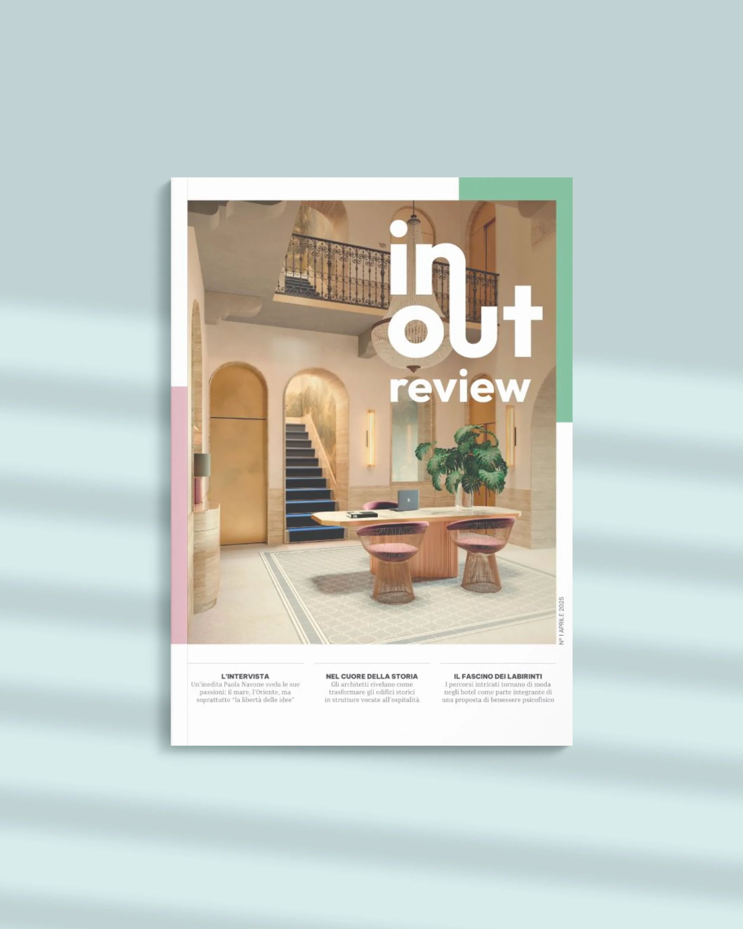 Copertina di una rivista intitolata 'In Out Review' con un'immagine di un interno elegante con scale, tavolo d'ufficio, sedie in tessuto, piante verdi e luci calde.