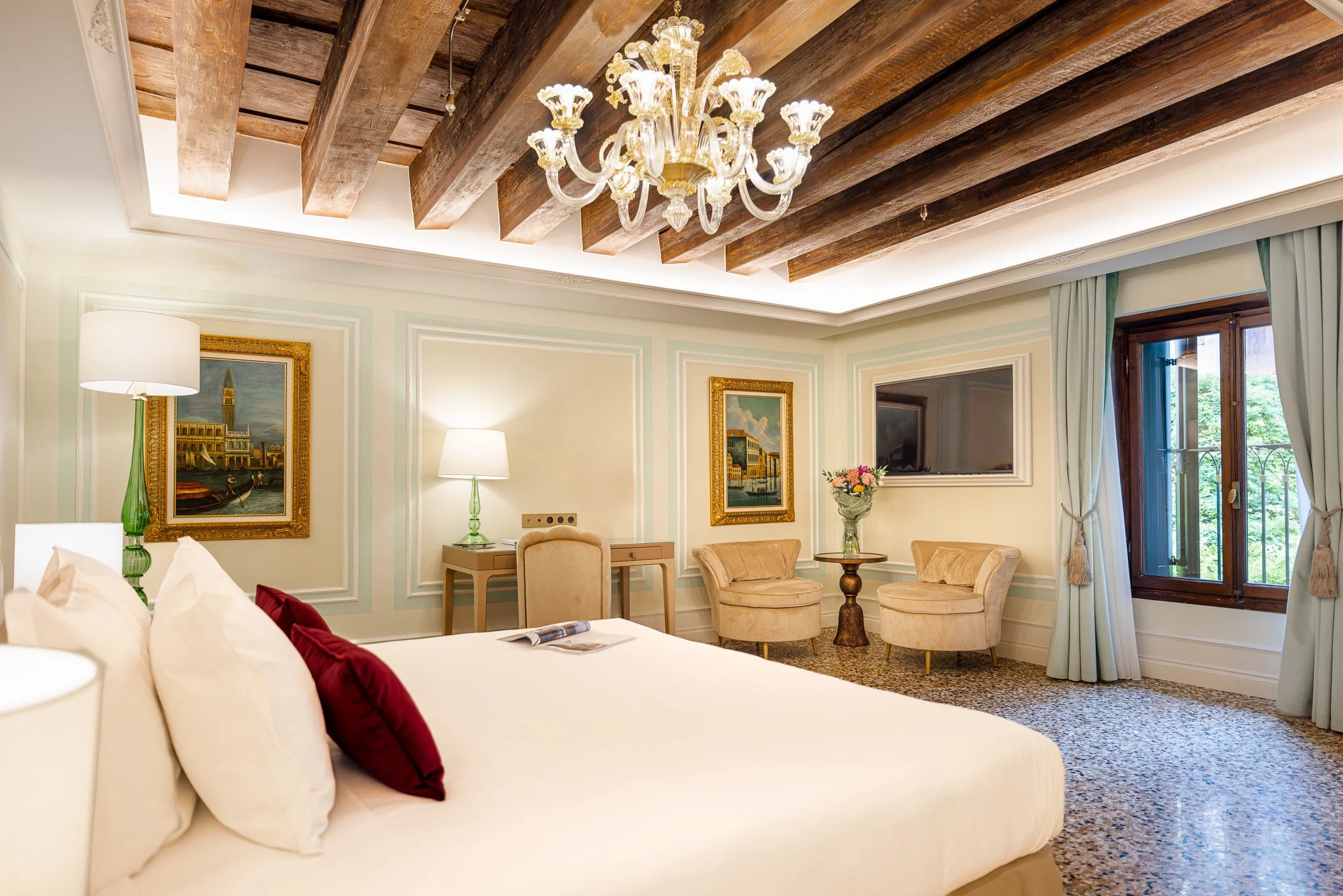 Camera da letto elegante con soffitto a travi in legno e lampadario di cristallo, mobilia raffinata, dipinti alle pareti, un grande finestrone con tende azzurre che si affaccia su un giardino verde.