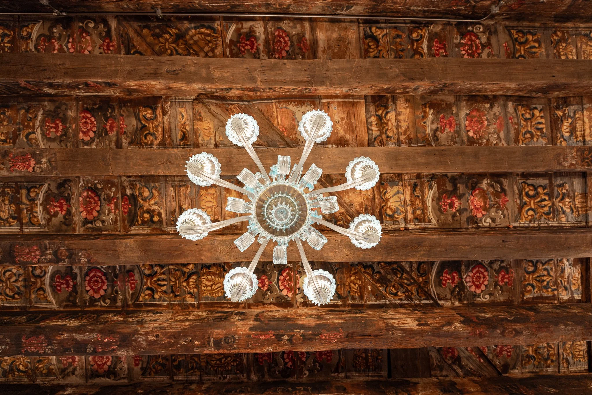 Lampadario di cristallo visto dal basso, appeso a un soffitto decorato con travi in legno e intarsi colorati.