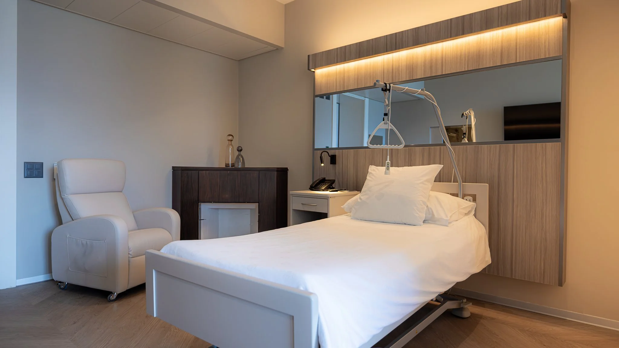 Camera d'ospedale con letto, sedia bianca, mobile scuro e specchio con luci a soffitto