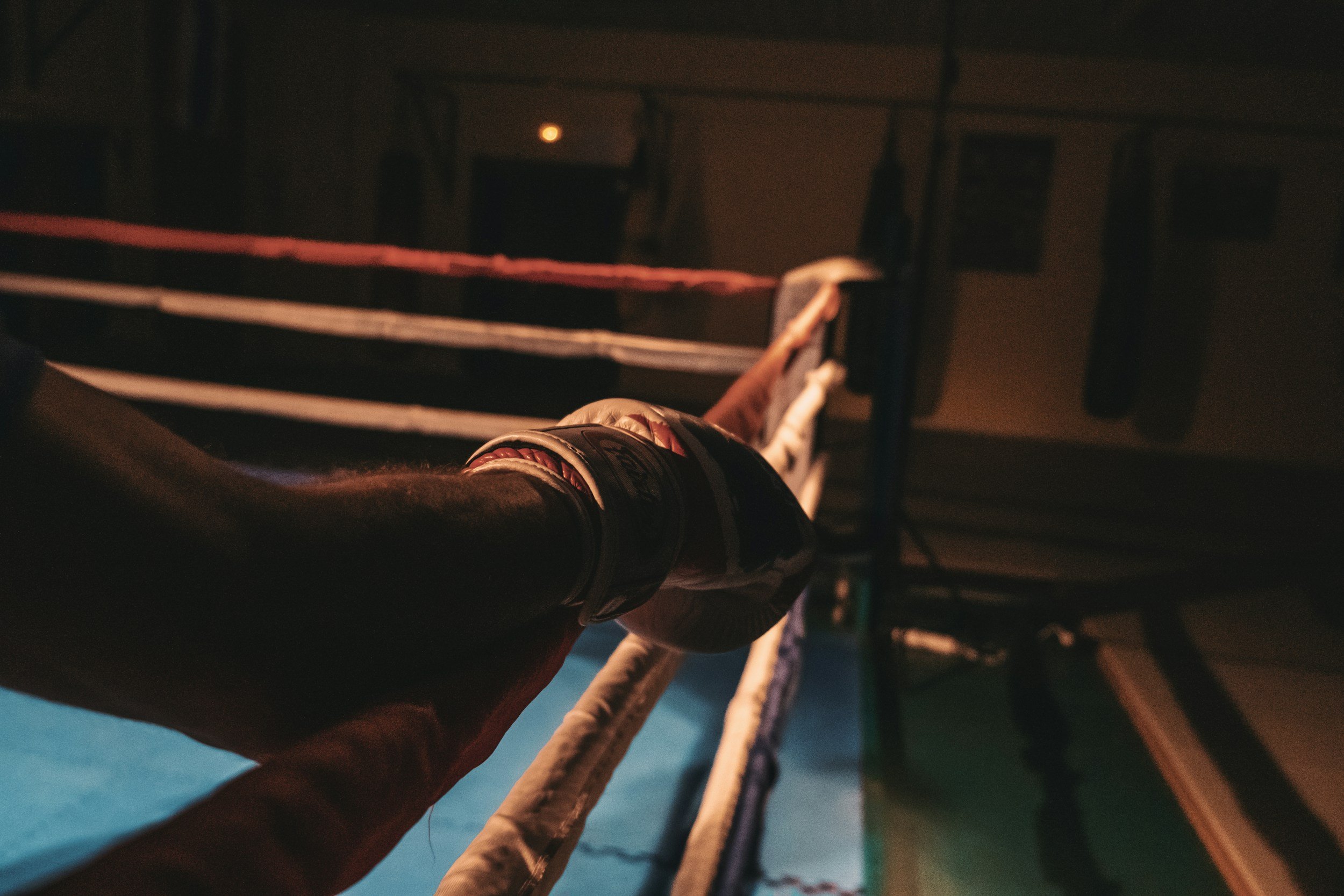 A boxer's gloved hand grips the corner ropes of a boxing ring, viewed from a low angle in a dimly lit gym.