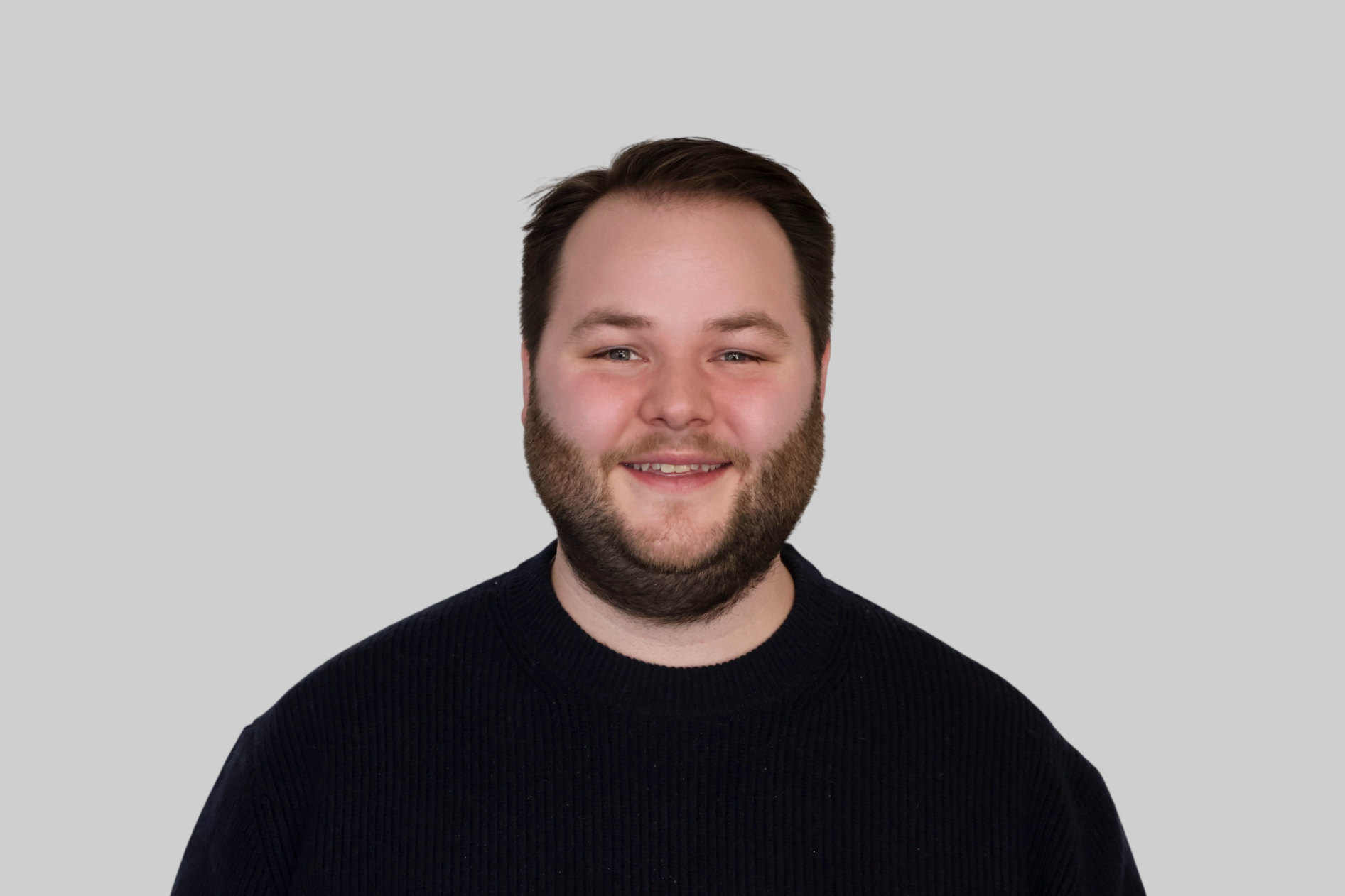 A smiling man with a beard and dark hair wearing a black sweater against a plain light gray background.