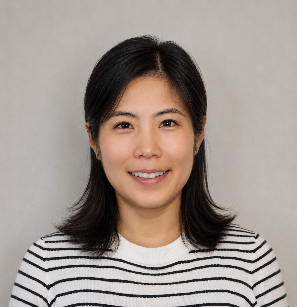 Portrait of a young woman with shoulder-length dark hair, smiling, wearing a white and black striped shirt, against a plain gray background.