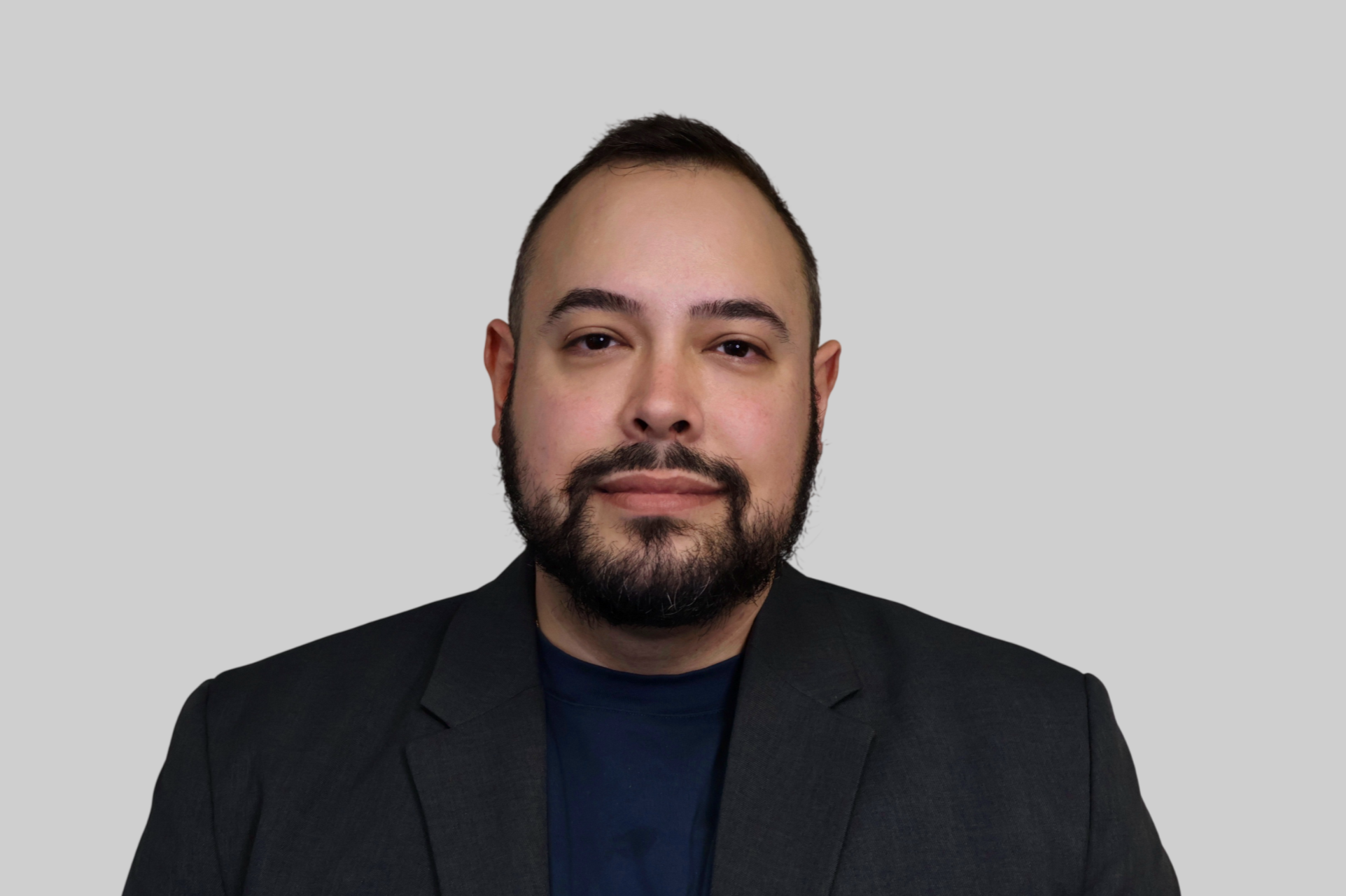 Portrait of a man with dark hair, beard, and mustache wearing a dark suit jacket and black shirt, against a plain light gray background.