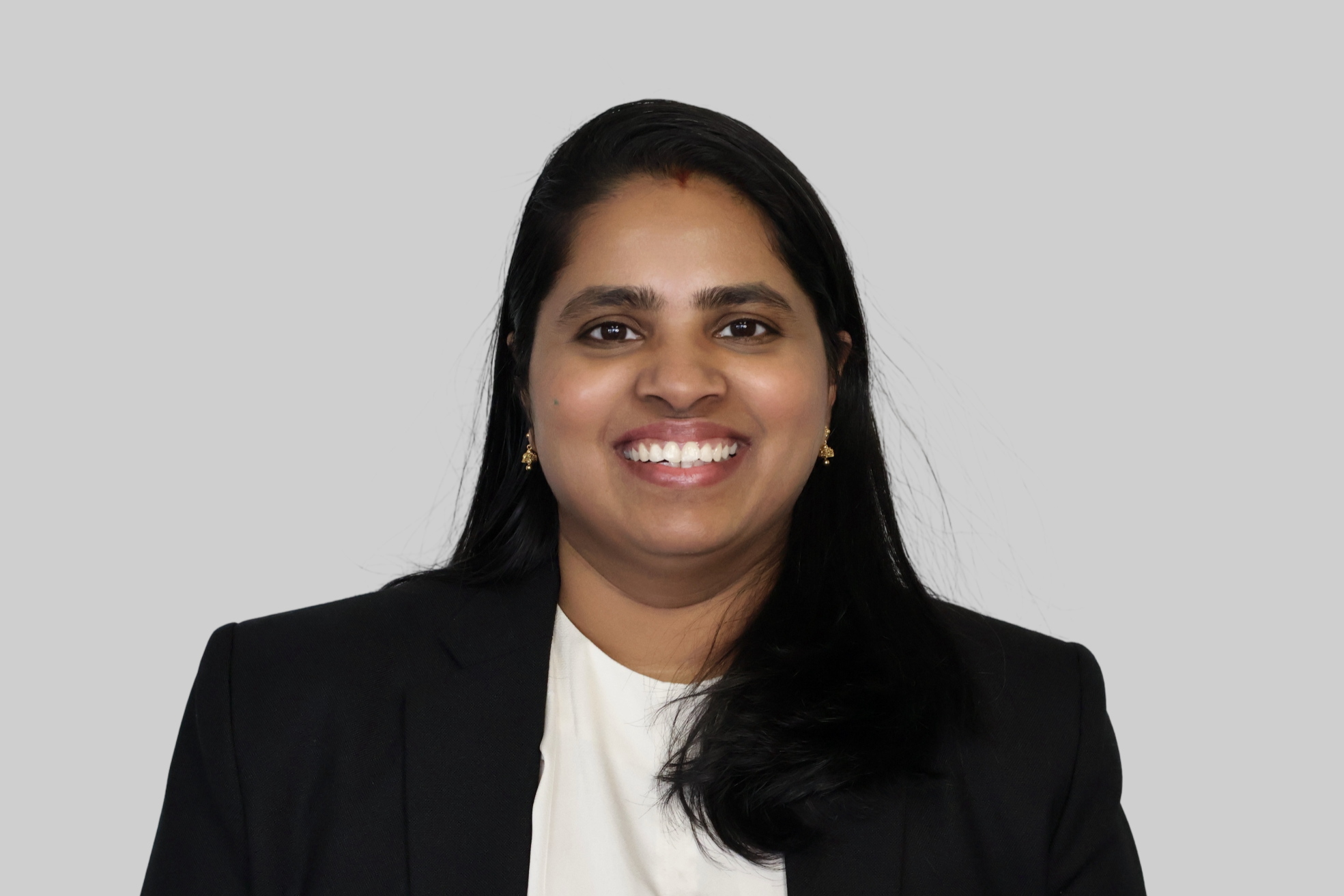 A woman with long black hair, dark eyes, and a bright smile, wearing a black blazer and gold earrings, posing against a plain gray background.