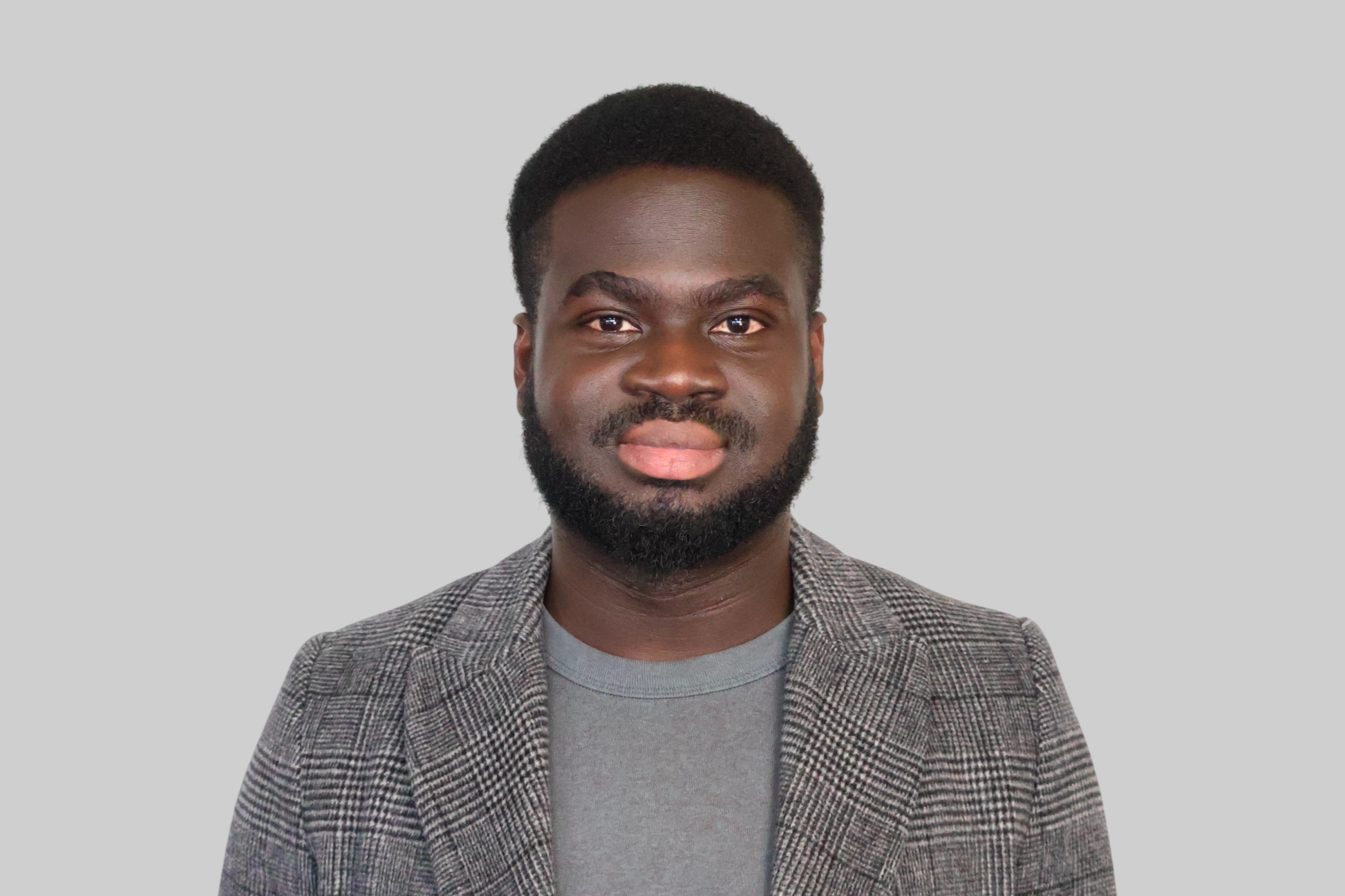 Headshot of a Black man with short curly hair and a beard, wearing a gray t-shirt and a plaid blazer, against a plain light gray background.