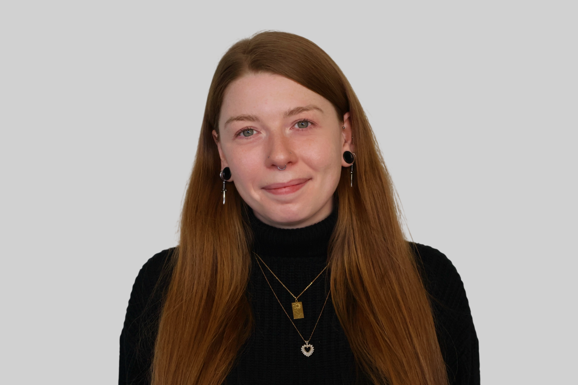 A young woman with long red hair, light skin, and multiple piercings, wearing a black turtleneck and layered necklaces, smiling slightly against a light gray background.