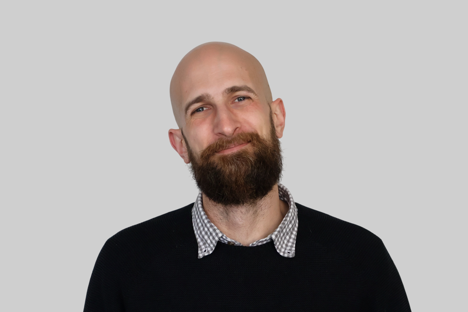 A man with a bald head and a full beard, smiling slightly, wearing a black sweater over a checkered shirt, standing against a plain light gray background.