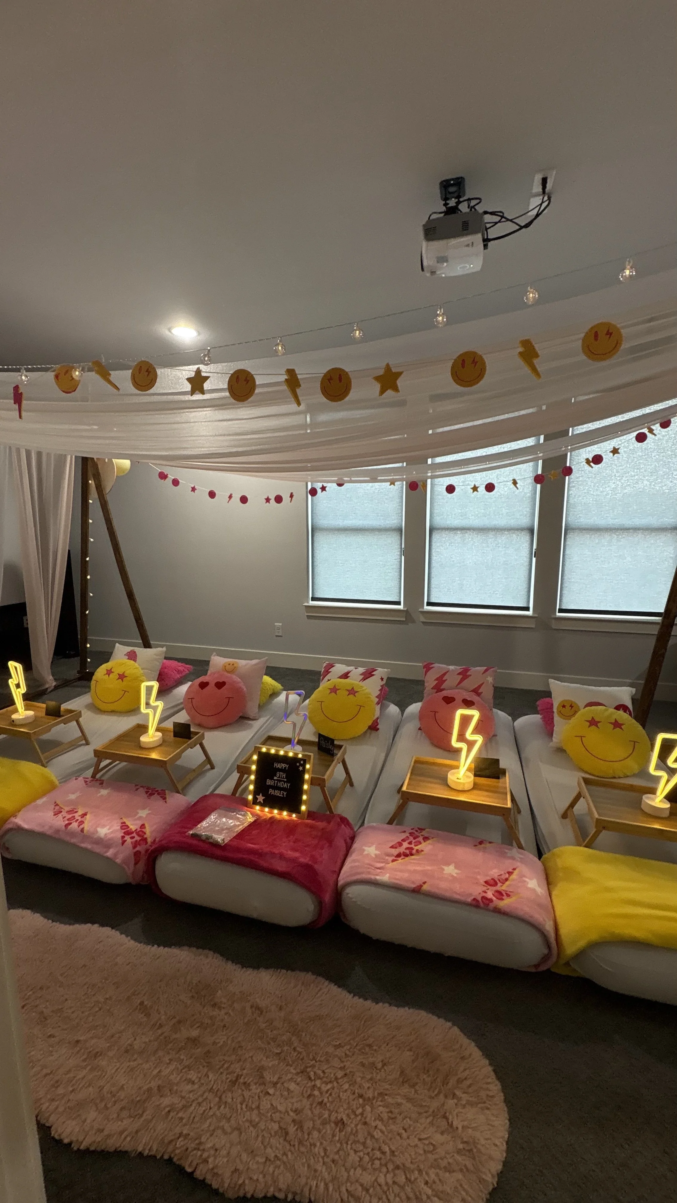 Decorated room with smiley face pillows, small tables with lightning bolt lamps, pink blankets, and a pink fluffy rug on the carpet. Hanging decorations include smiley face, star, and lightning bolt shapes. The room appears to be set up for a birthda