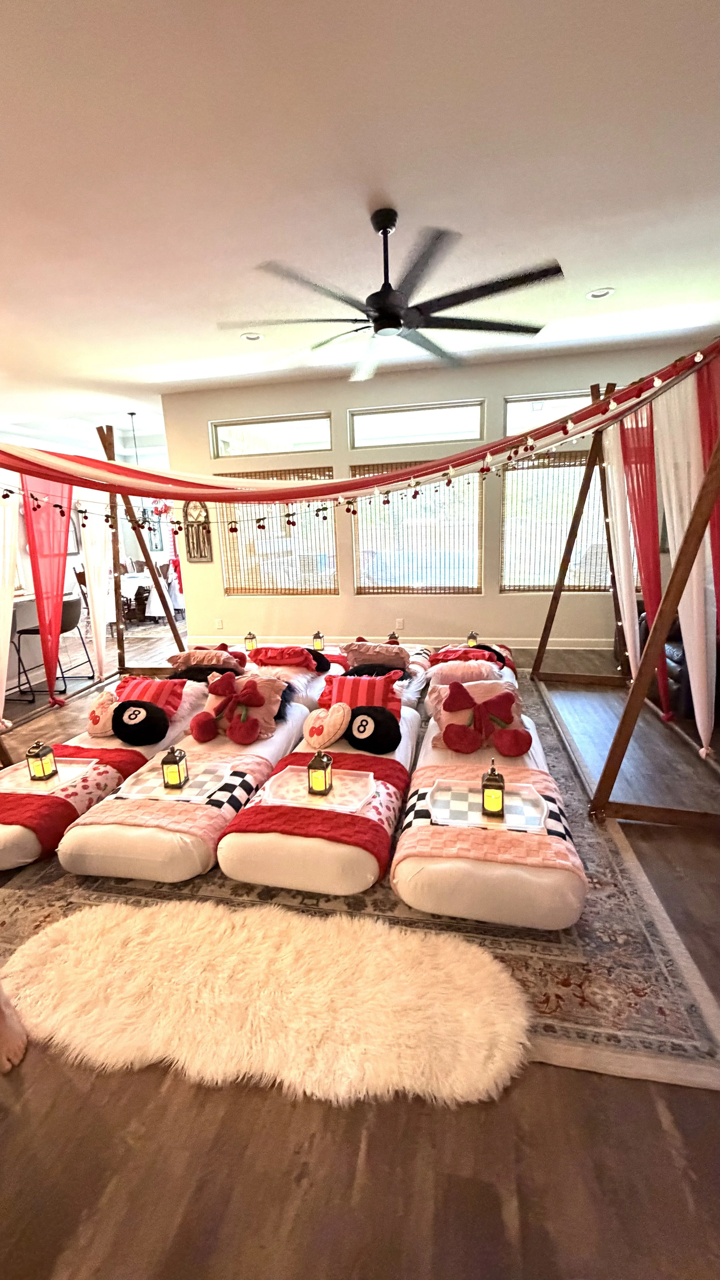 Indoor cozy setup with multiple mattresses, pillows, and blankets in red and pink hues, decorated with plush cherry and heart pillows, surrounded by lanterns, with a fluffy white rug on a wooden floor, and a swing with sheer red and white curtains overhead.