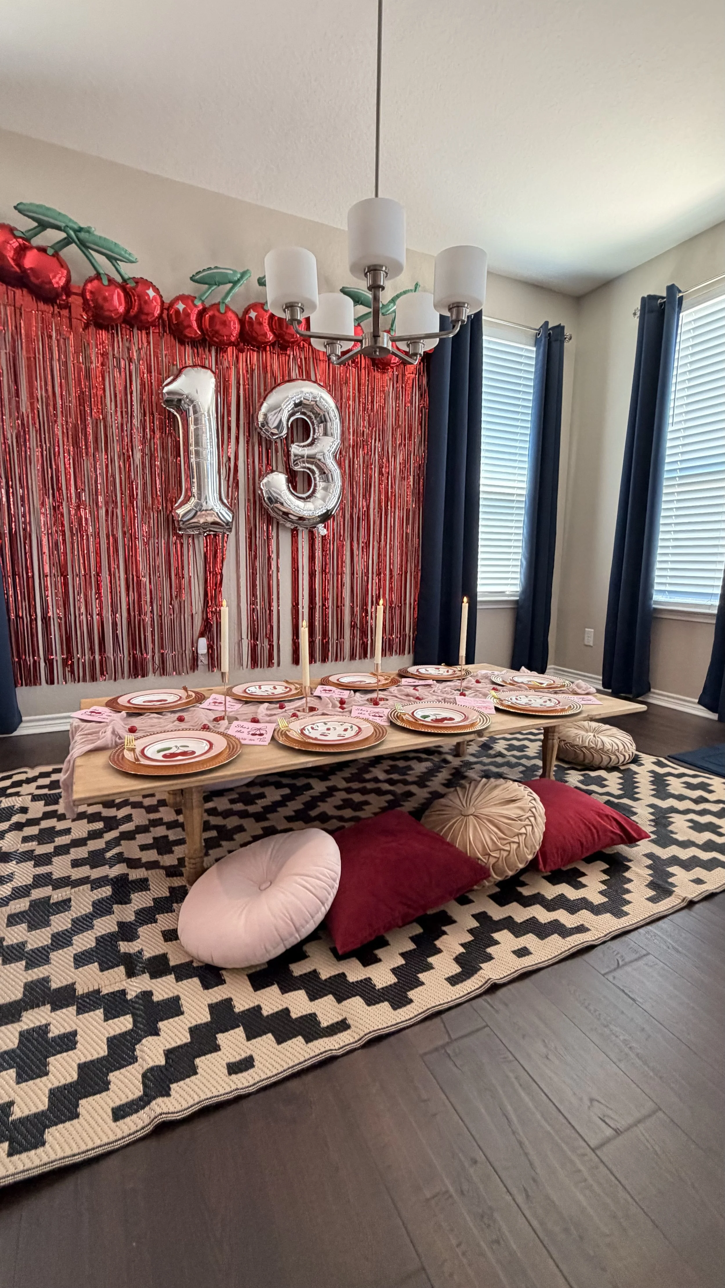 A low table set up for a meal with paper plates, pink napkins, and pink and gold utensils, on a patterned black and beige rug. Cushions in beige, red, and pink are arranged around the table. Behind the table, a red curtain with metallic foil fringe a
