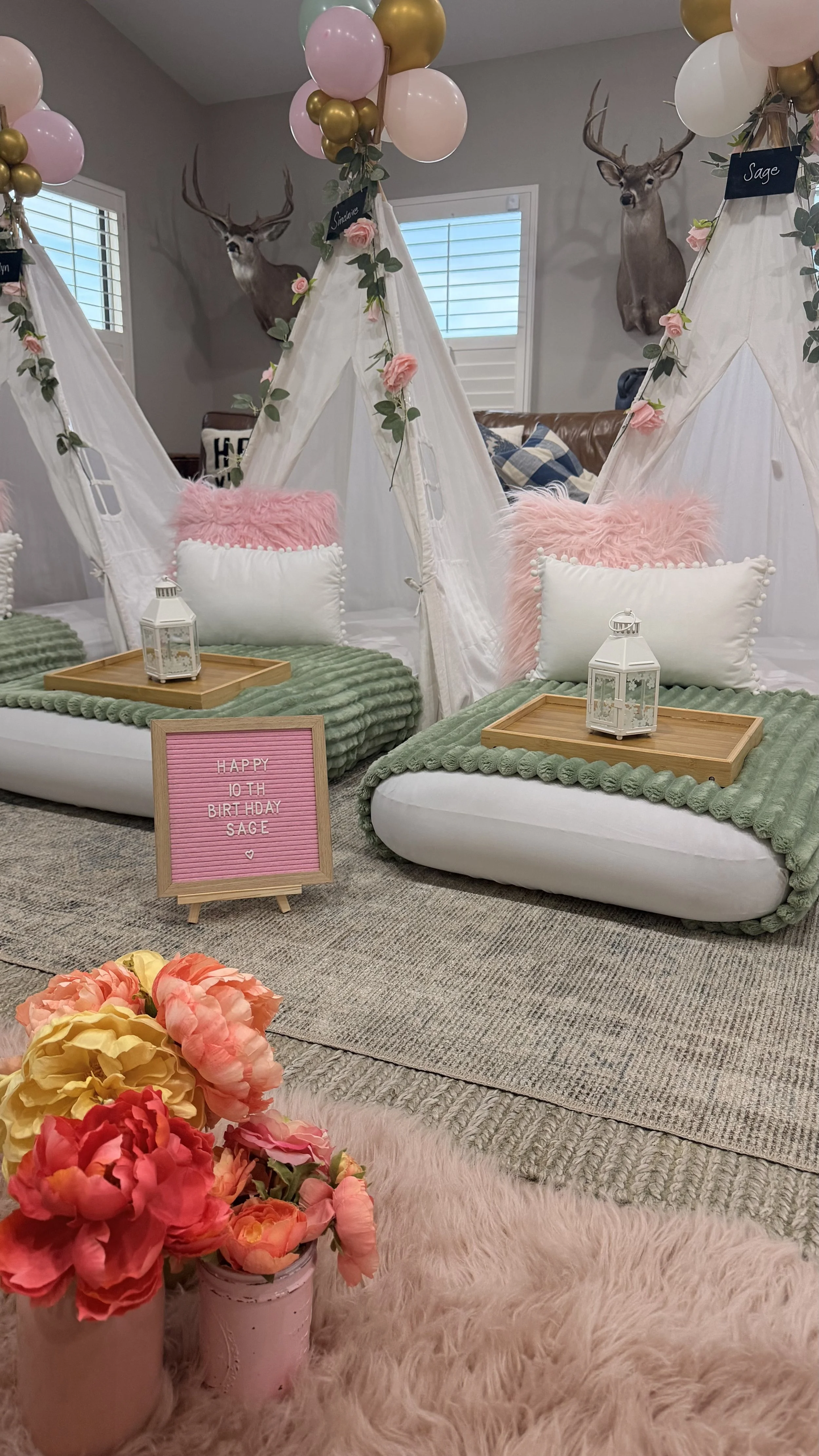 Decorative indoor setting with two small white tepees draped in white fabric, decorated with pink roses and green leaves, each with fluffy pink and white pillows and wooden trays with lanterns on top. A pink sign reads 'Happy 10th Birthday Sage.' Pin