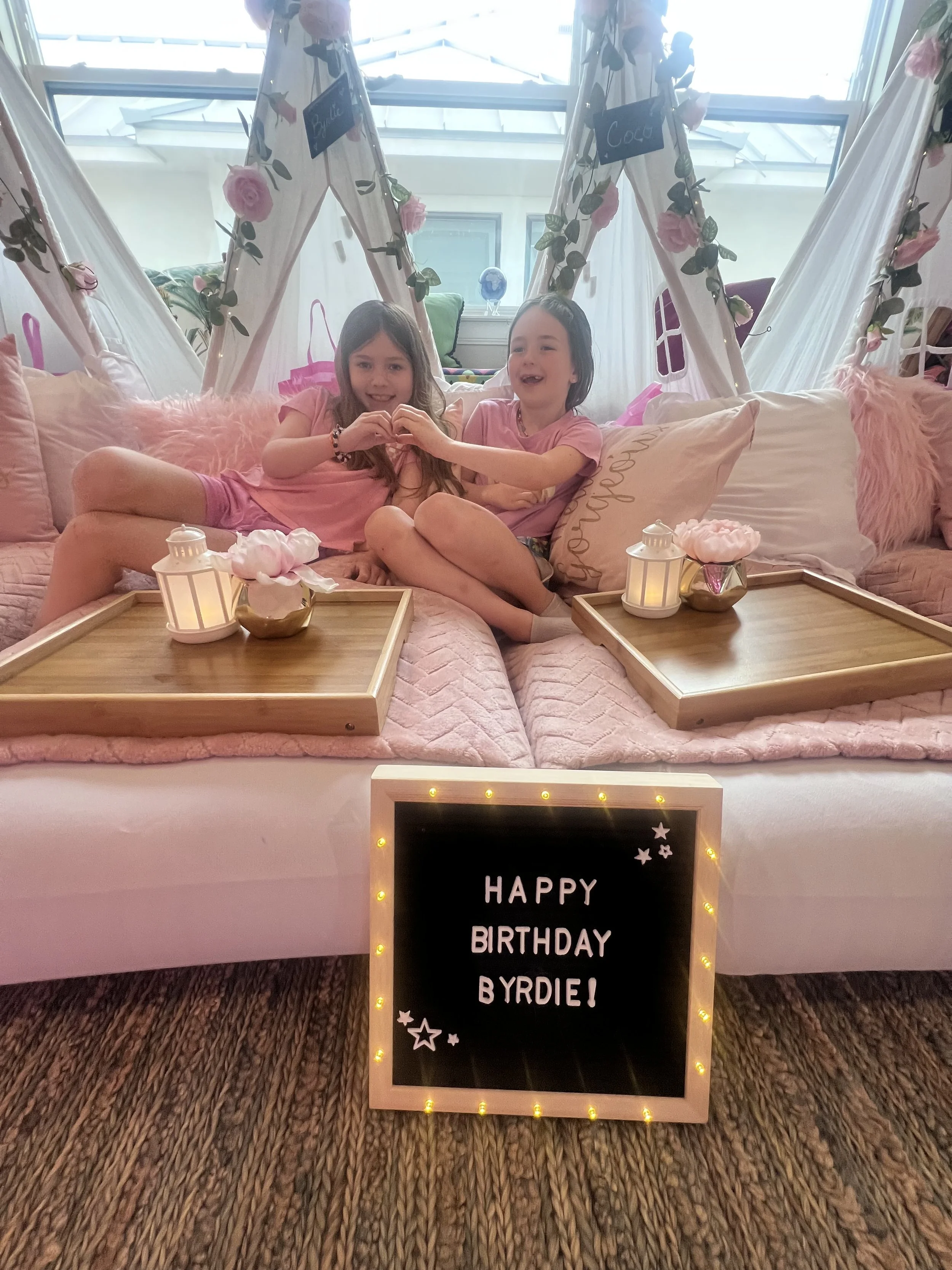 Two young girls sitting on a bed with pink pillows and blankets, forming a heart shape with their hands. The background features a canopy tent decorated with pink roses and fairy lights, and a window with a globe on the windowsill. There is a signboa