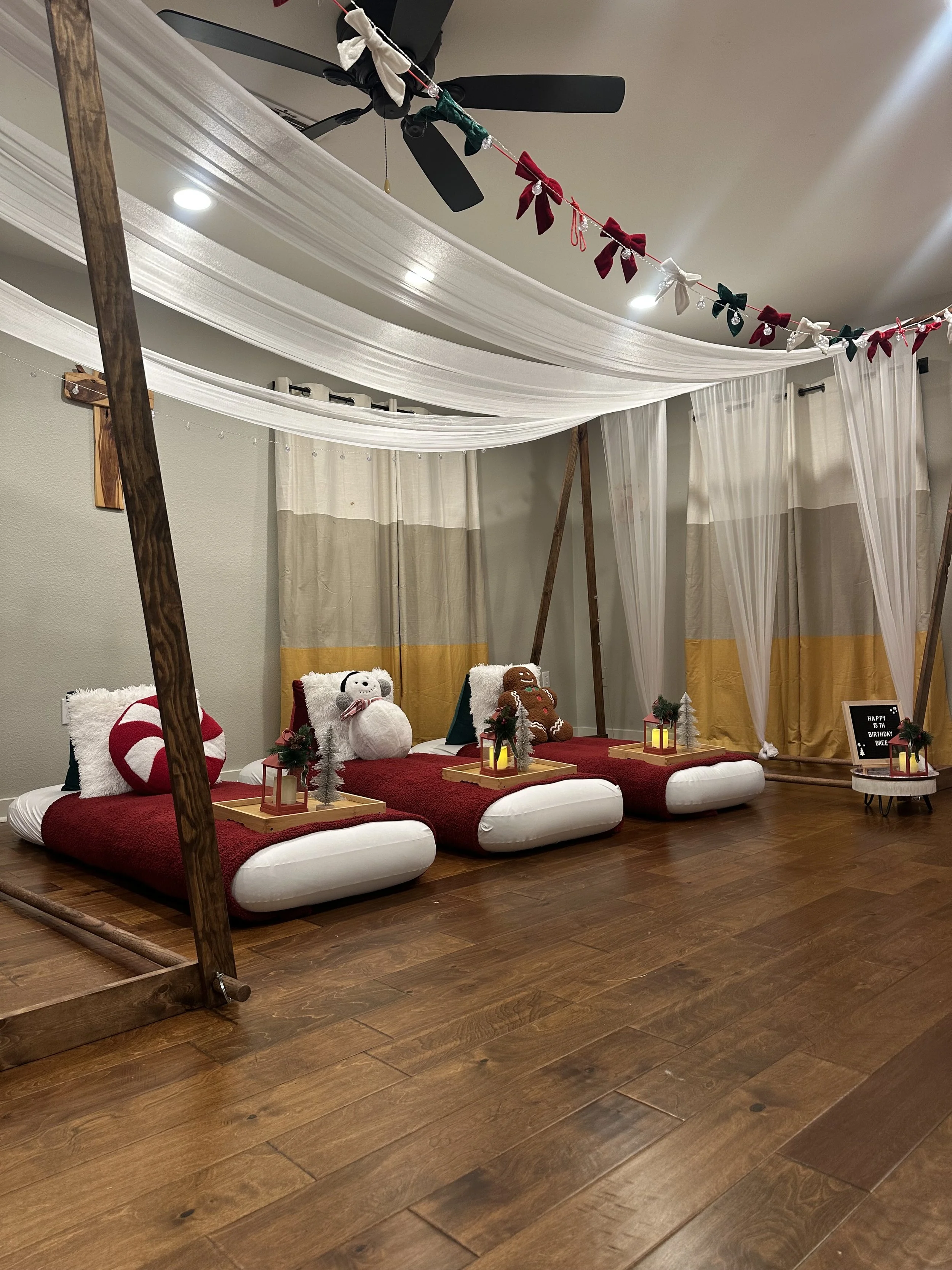 Indoor setup with traditional Japanese seating on red cushions, decorated for Christmas with plush toys, lanterns, and garlands, under draped white fabric and curtains, with a wooden frame and ceiling fan.
