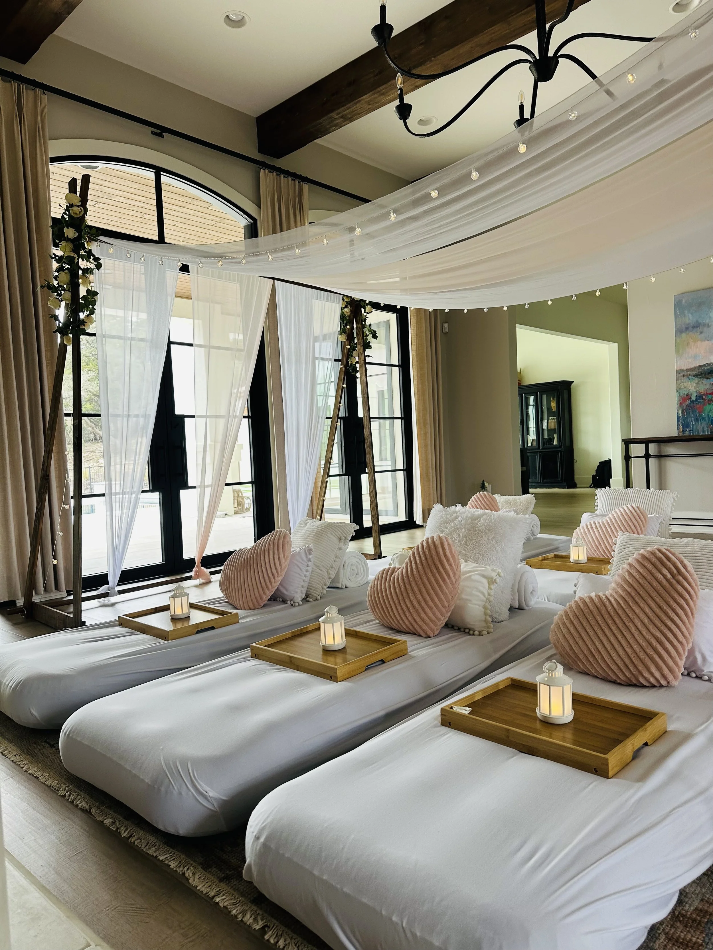 A cozy indoor space decorated for a celebration or event, featuring large beds with white linens, pink heart-shaped pillows, wooden trays with candles, large windows with sheer white curtains, and a chandelier hanging from the ceiling.