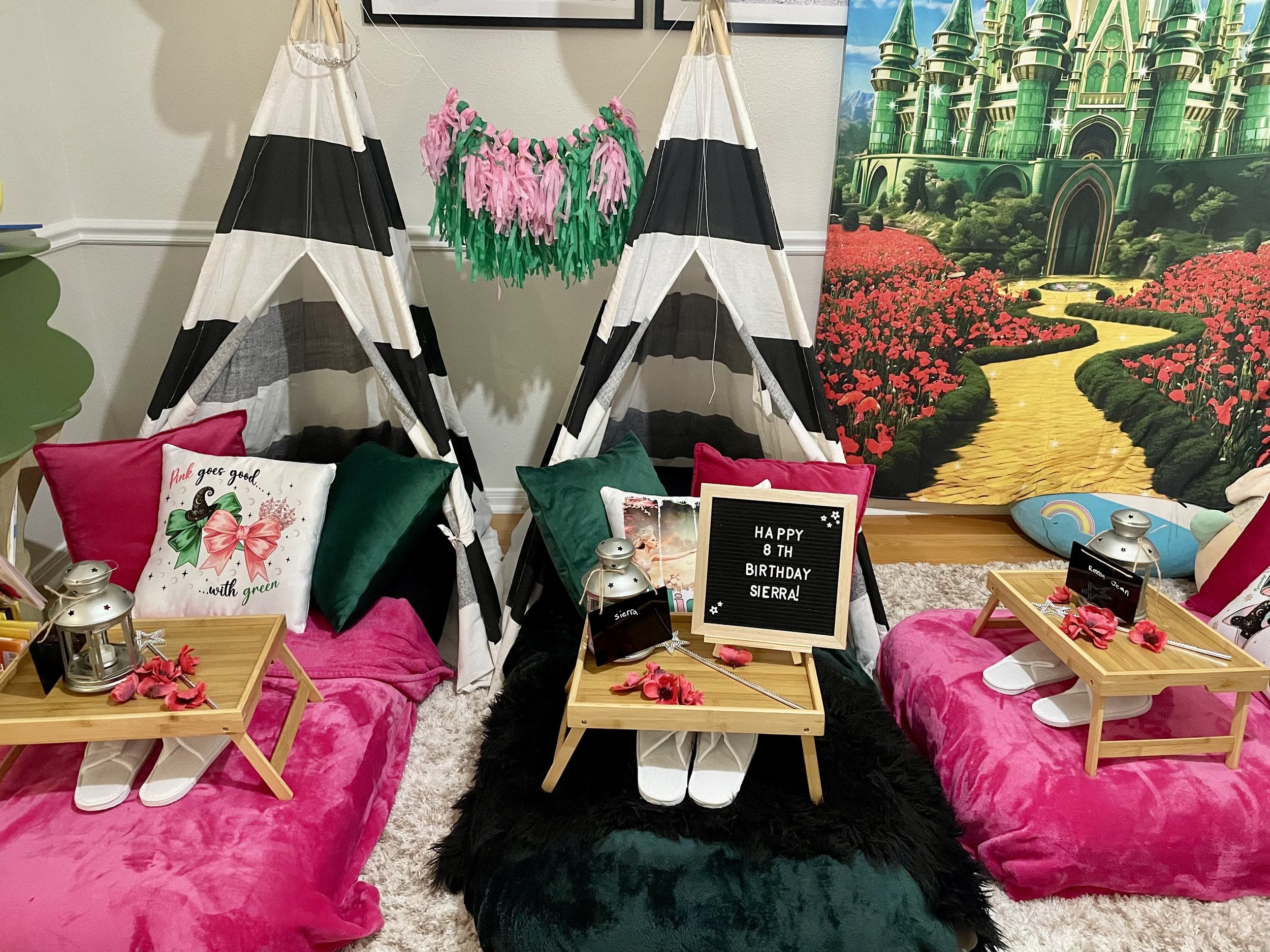 Decorated indoor scene with pink and green pillows, small wooden trays, lanterns, and pink flower petals on soft pink cushions. There is a teepee, a black faux fur rug, a Hogwarts castle poster, and various birthday decorations including a blackboard
