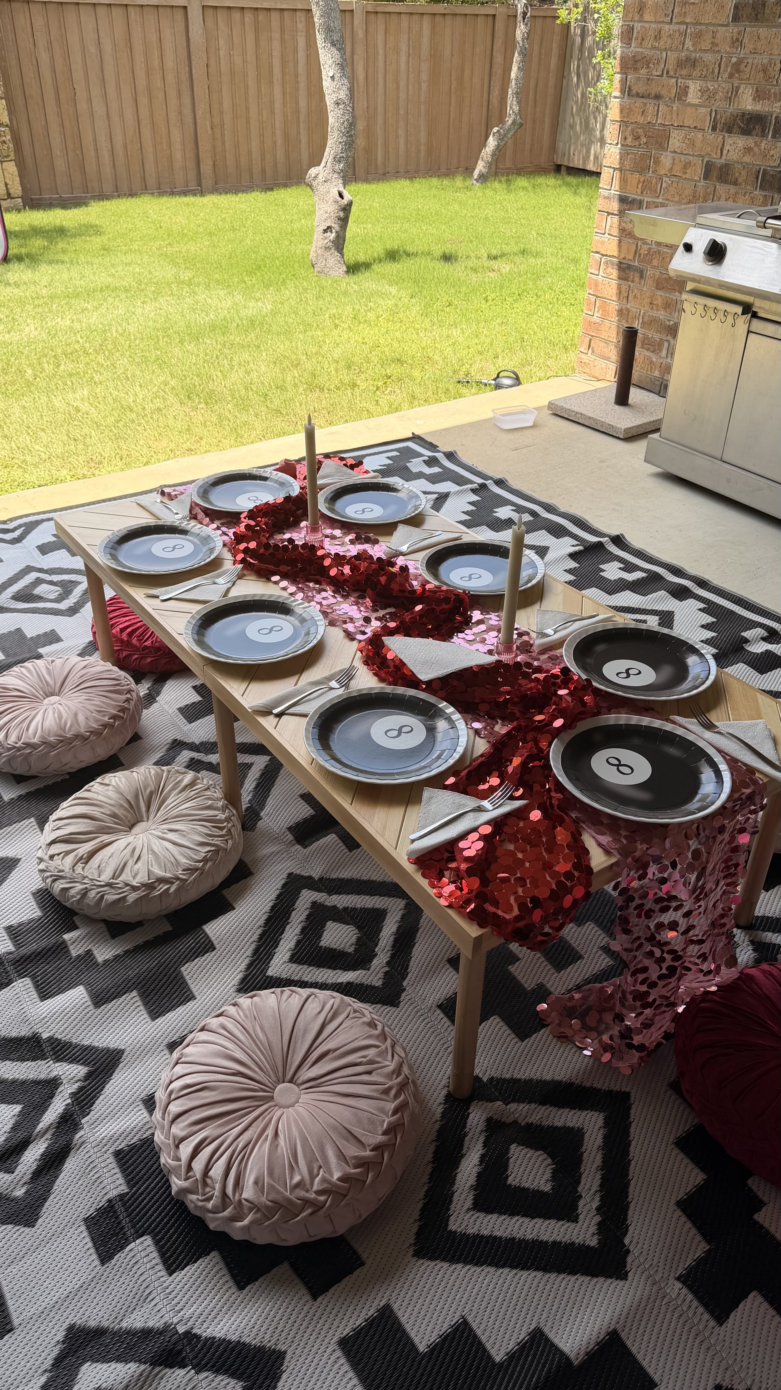 Outdoor dining table setup for a party on a covered patio. The table has black and white place settings with the number 8 on each plate, a red sequin table runner, and two white candles. Several round pink cushions are on the patterned rug surroundin
