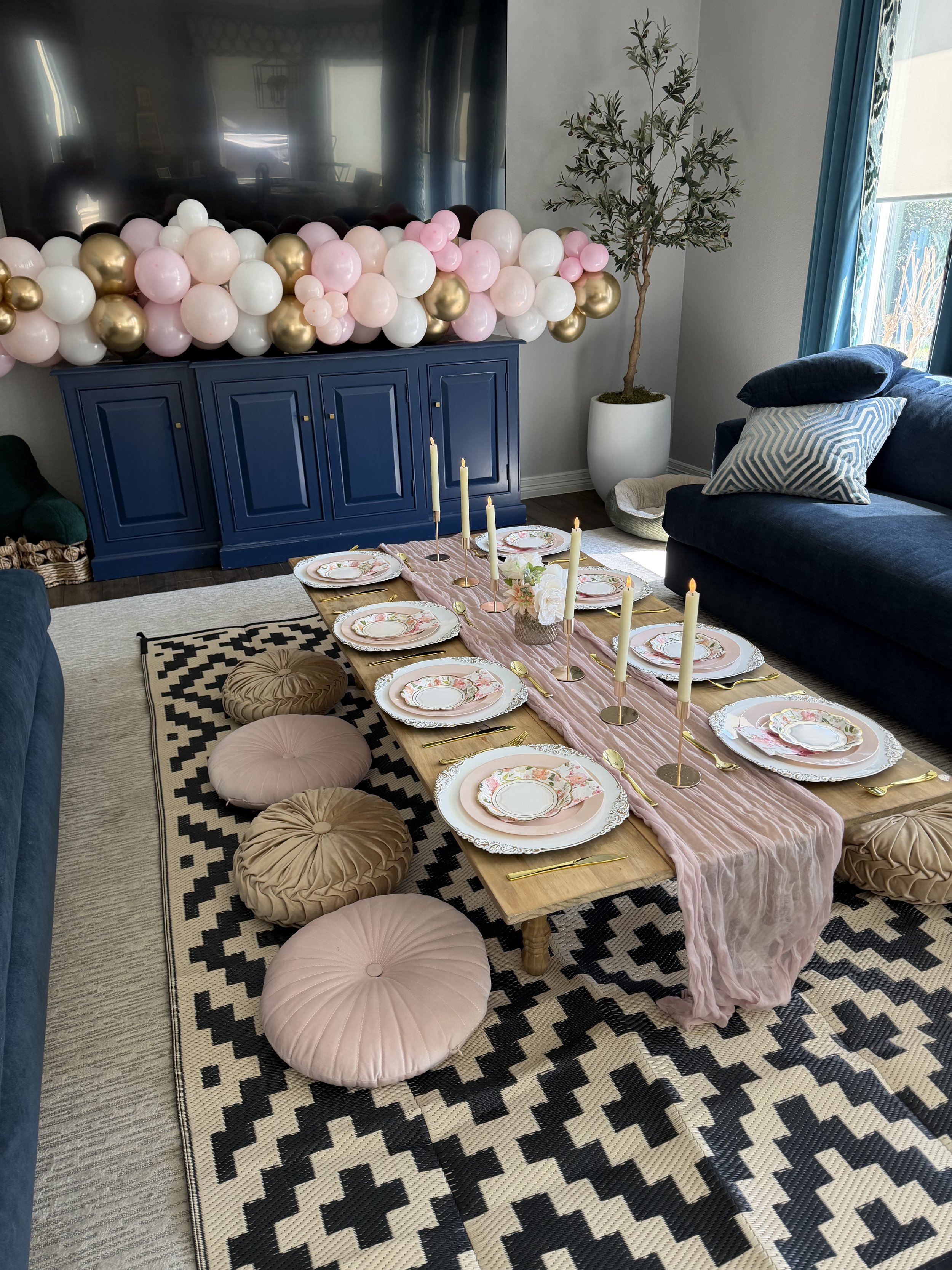 Living room decorated for a celebration with a dining table set with floral plates, gold utensils, and pink candles, pink, white, and gold balloons arranged over a blue cabinet, a potted olive tree, and a black and white patterned rug on the floor.