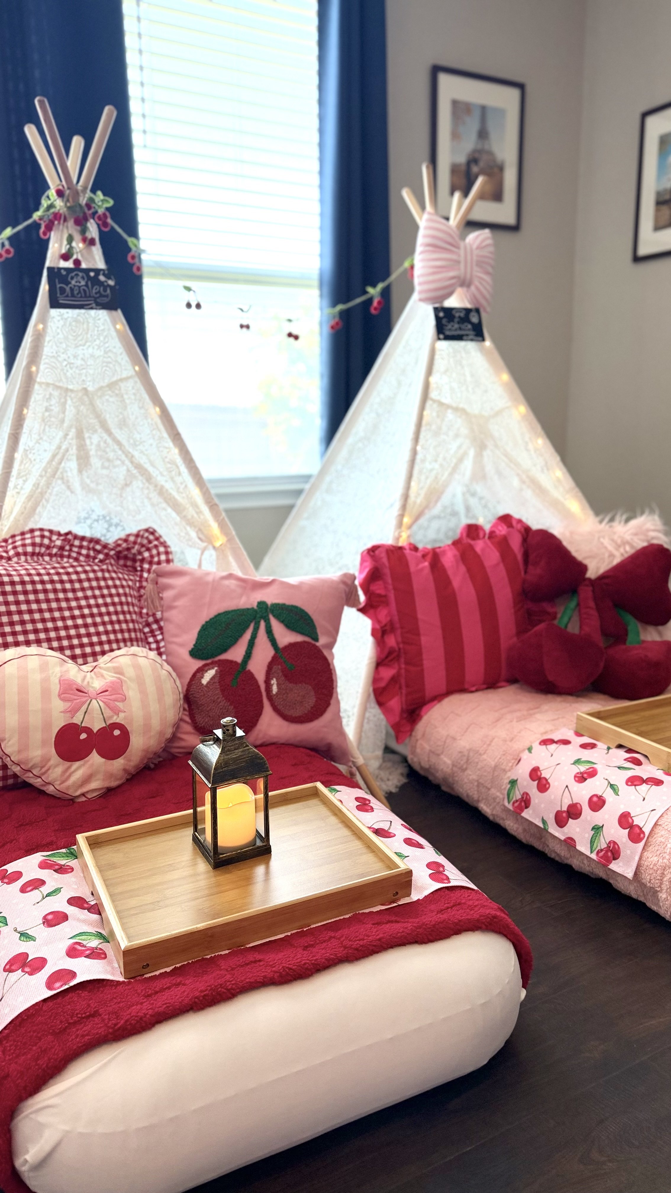 Indoor scene featuring two white tipi tents decorated with string lights, set up in a room with dark hardwood floors, framed pictures of the Eiffel Tower on the wall, and blue curtains. The area is decorated with cherry-themed cushions, pillows resem