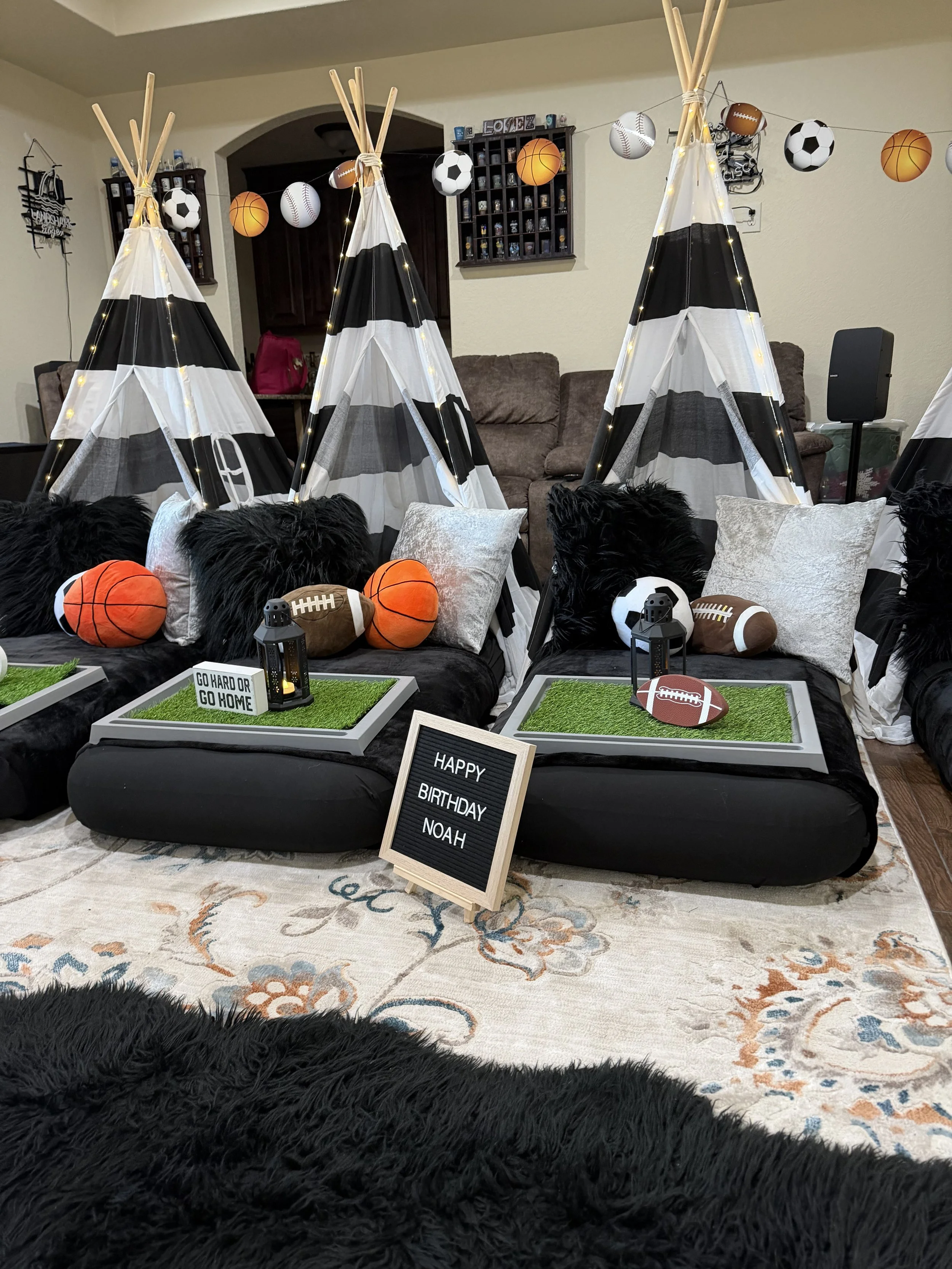 Indoor sports-themed birthday party setup with black and white teepees, plush pillows, sports balls, string lights, lanterns, and signs including a chalkboard sign that reads "HAPPY BIRTHDAY NOAH".