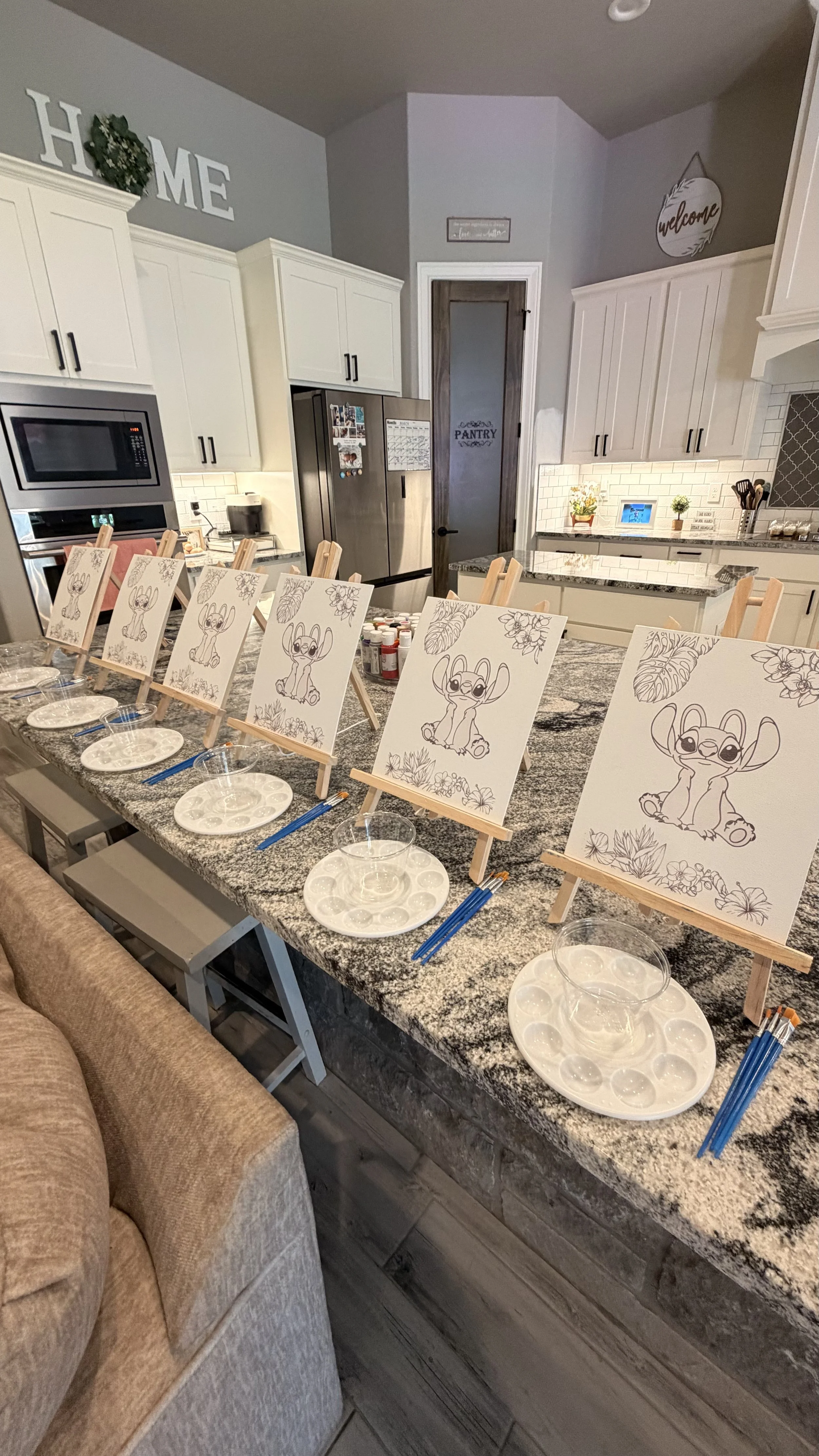 A kitchen with a long granite island counter, decorated with set up for a coloring activity featuring cartoon characters on small easels, with plates, water cups, and paint palettes. The kitchen has white cabinets, a gray backsplash, and decorative s
