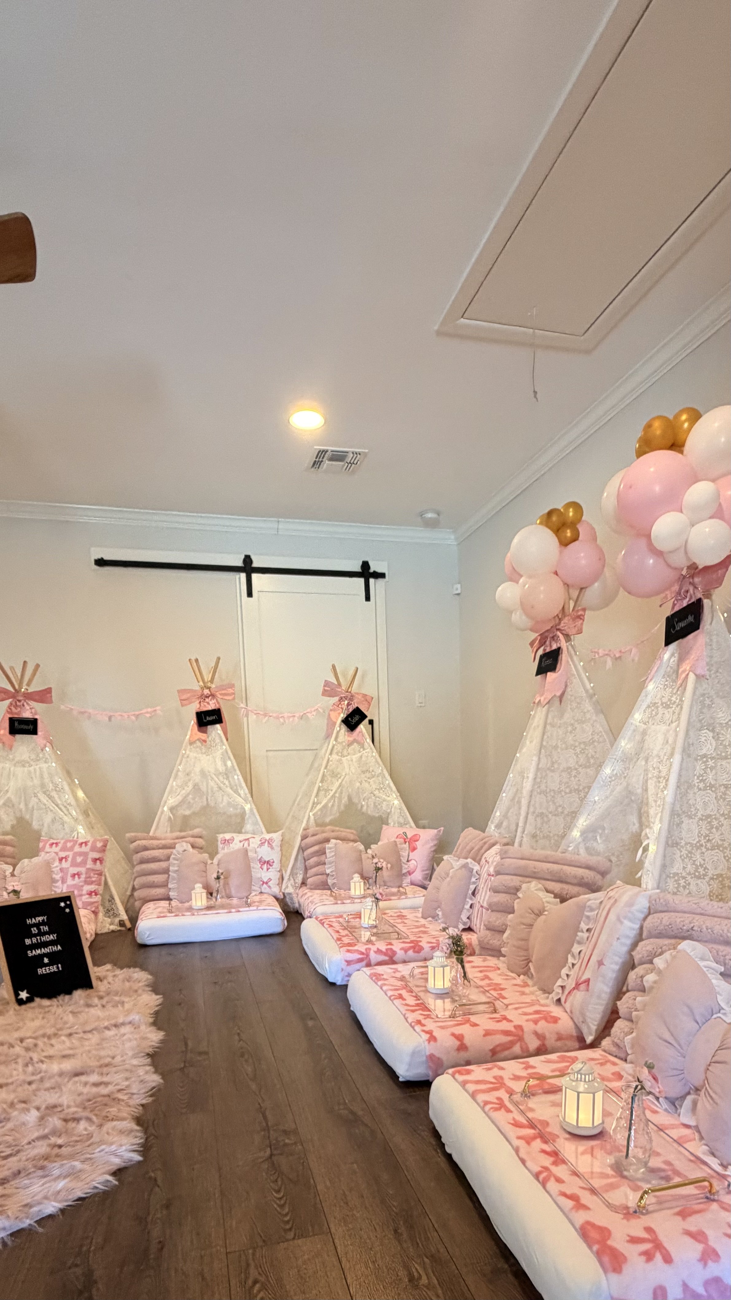 Decorative indoor space prepared for a birthday party with pink, white, and gold color theme. There are teepees, pillows, candles, balloons, and a blackboard sign that reads 'Happy 13th Birthday Samantha & Reese!'.