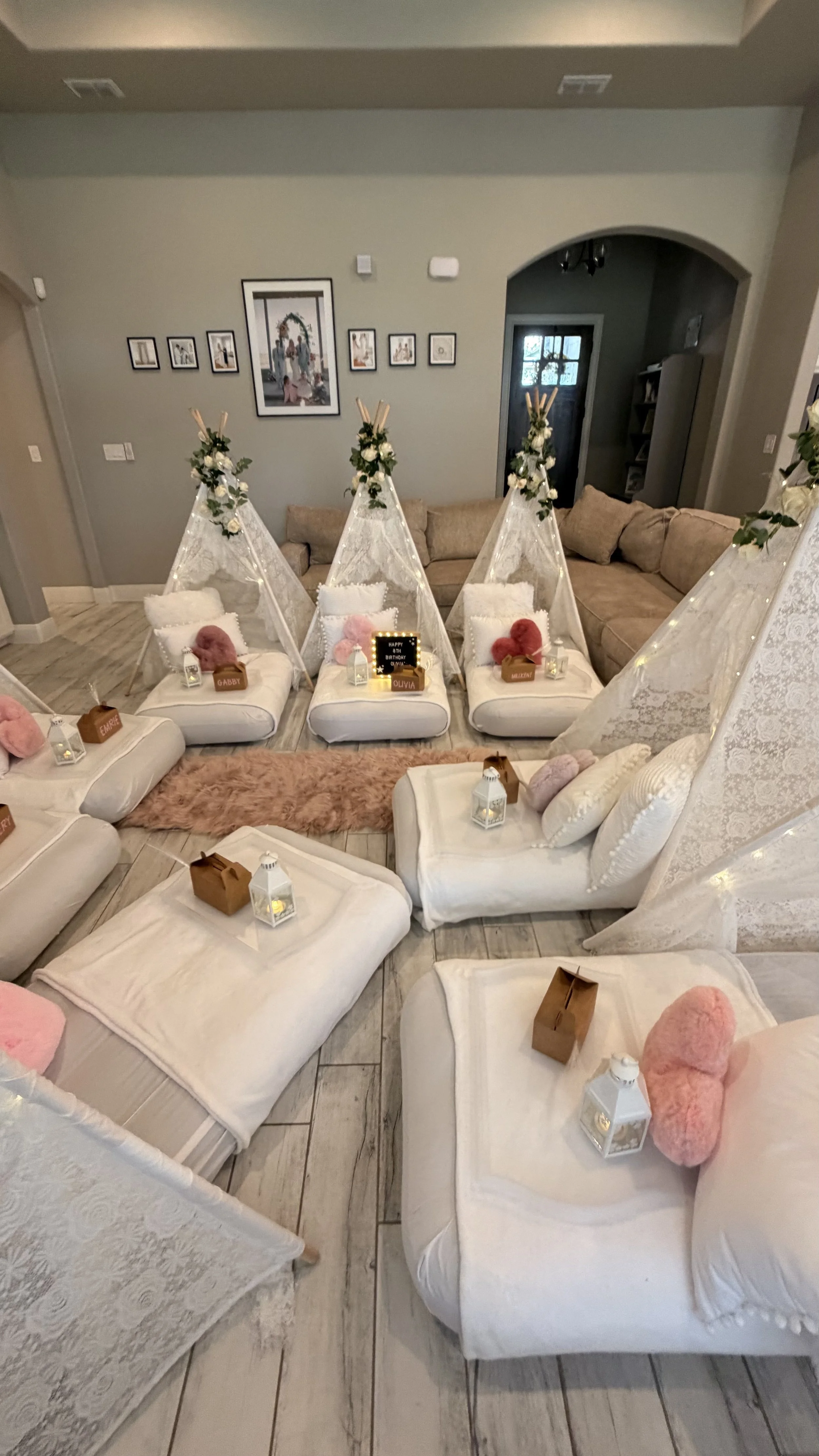 A cozy living room decorated for a celebration with teepee tents, plush pillows, lanterns, and stuffed teddy bears, in addition to a beige sectional sofa and wall art.