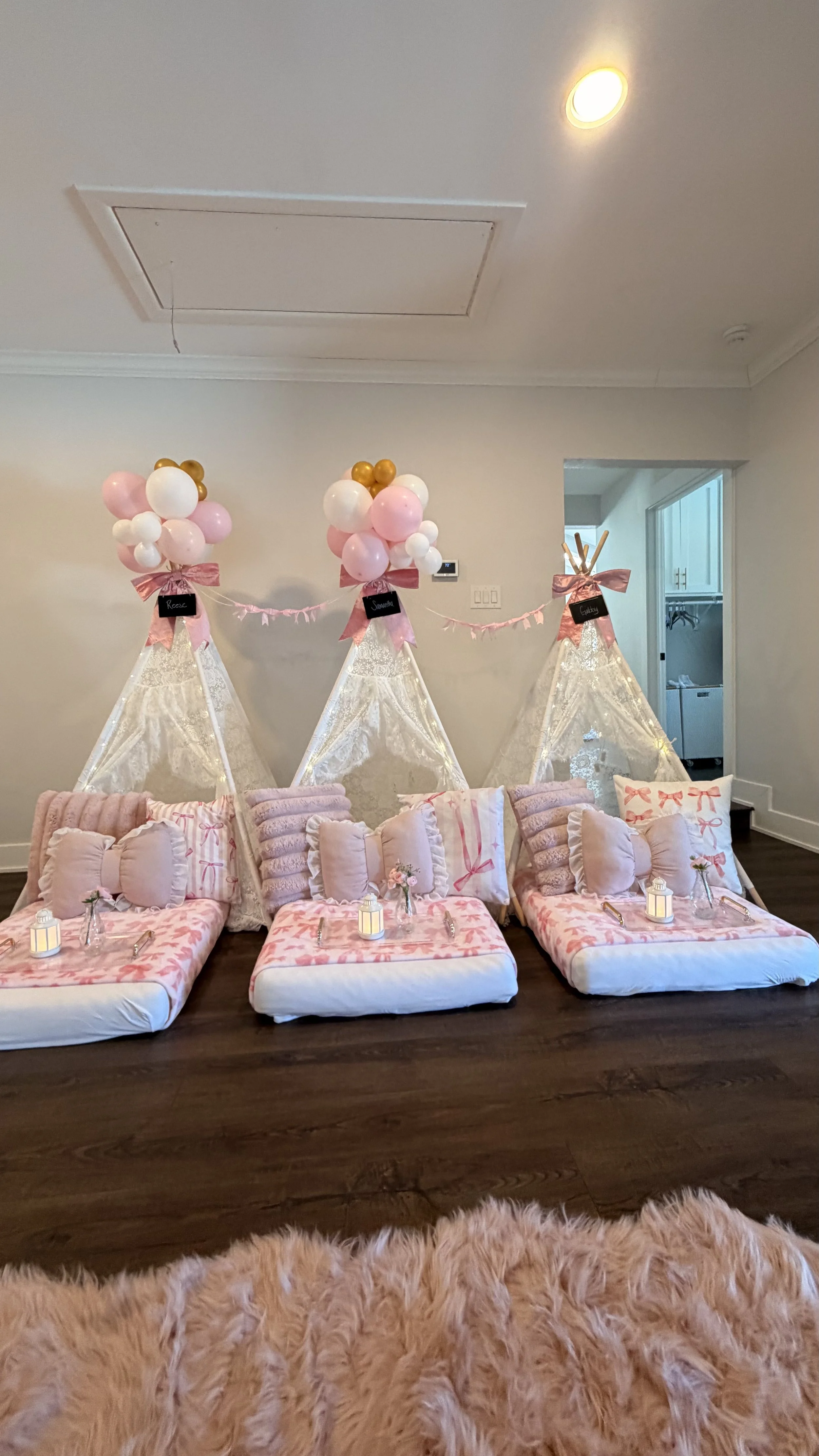 A cozy indoor space decorated for a celebration or gathering, featuring three small beds with pink and white pillows and blankets, set against a wall with lace teepees topped with pink and gold balloons and pink bows. Small lanterns are placed on the
