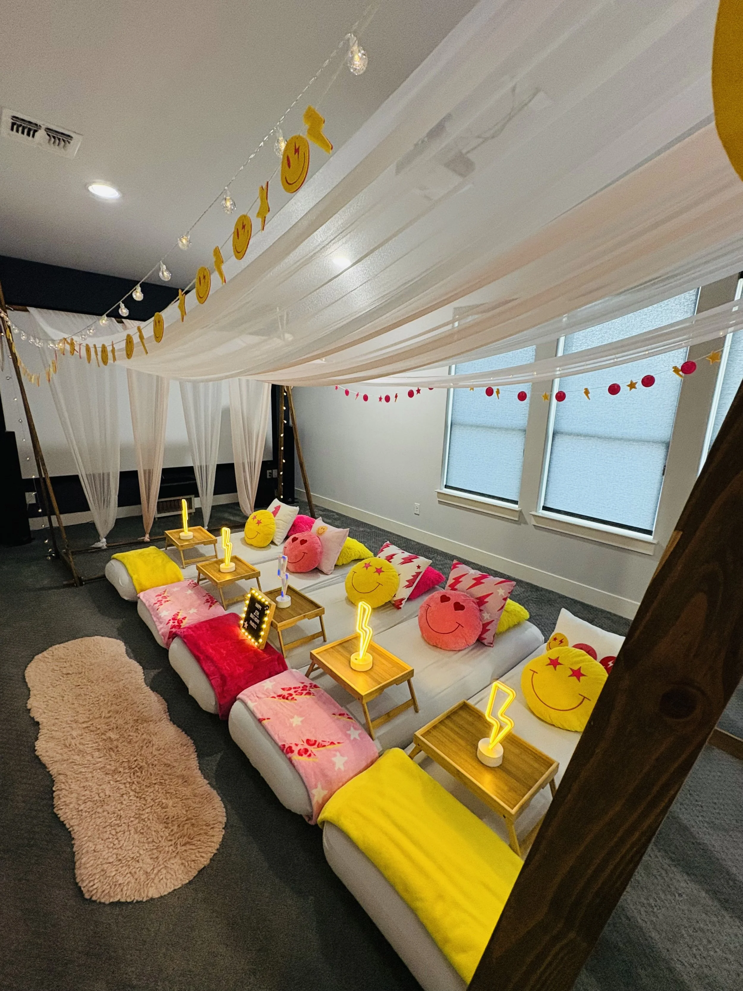Indoor lounge area decorated with smiley face and star-themed cushions, pink and yellow blankets, small wooden tables with illuminated lightning bolt decor, pink fluffy rug, sheer white drapes, string lights, and red and yellow hanging garlands.