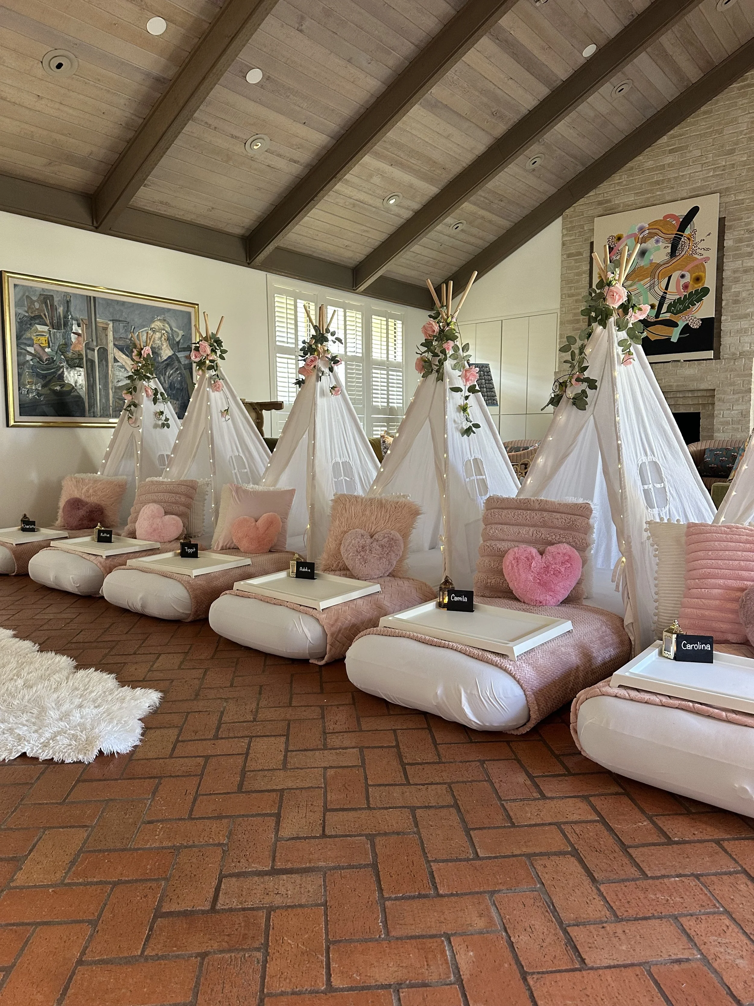 Row of small white couches with pink cushions shaped like hearts, decorated with flower garlands, in a living room with brick floor, large painting, and white walls.