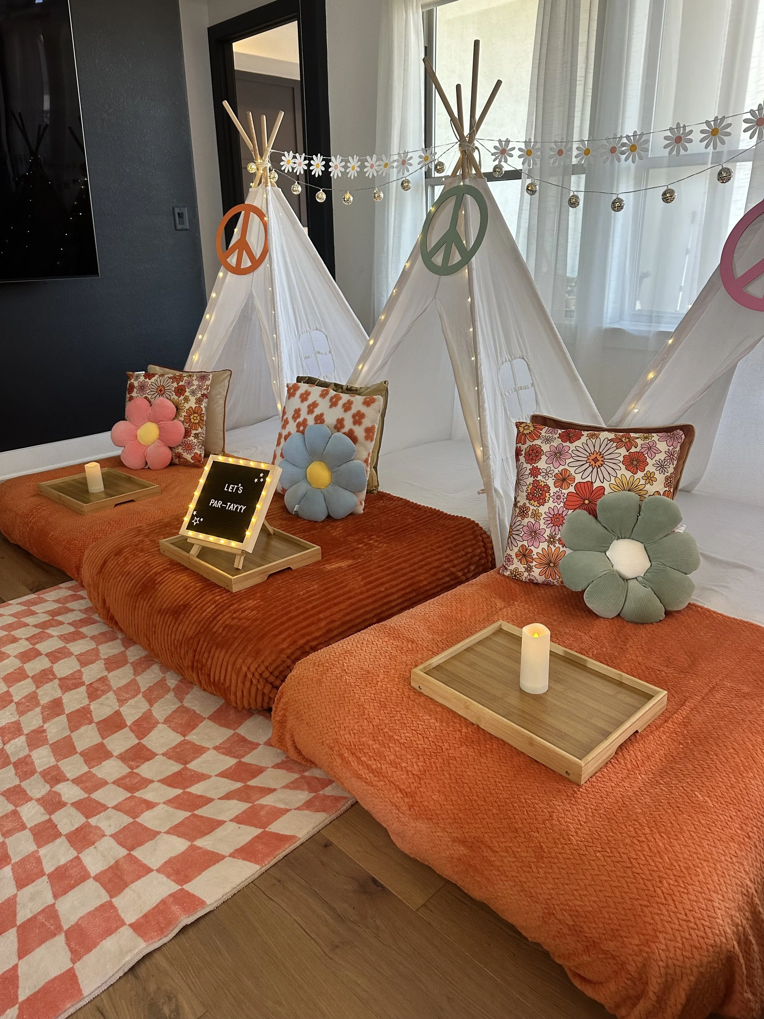 Cozy indoor scene with two low beds covered with orange blankets, decorated with floral pillows and plush flower cushions. Small wooden trays with candles and a sign reading 'Let's Par-Tayyy' are on the beds. Tepees with peace symbols, fairy lights, 