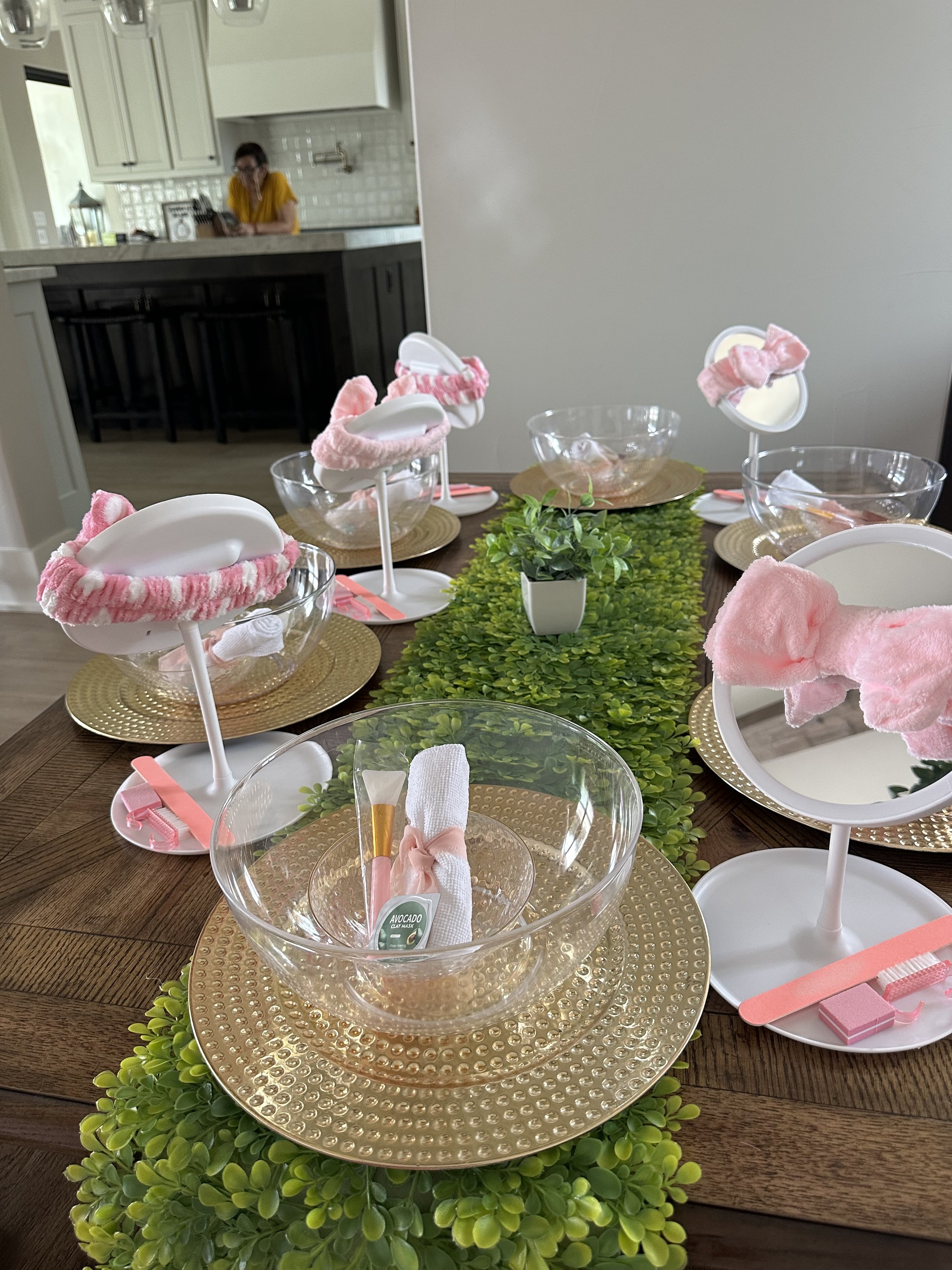 A dining table is set with clear glass bowls, pink plush eyemasks, and utensils wrapped in white cloth with pink ribbons, seated on gold chargers, with a green leafy table runner. In the background, a kitchen with white cabinets and a woman in a yell