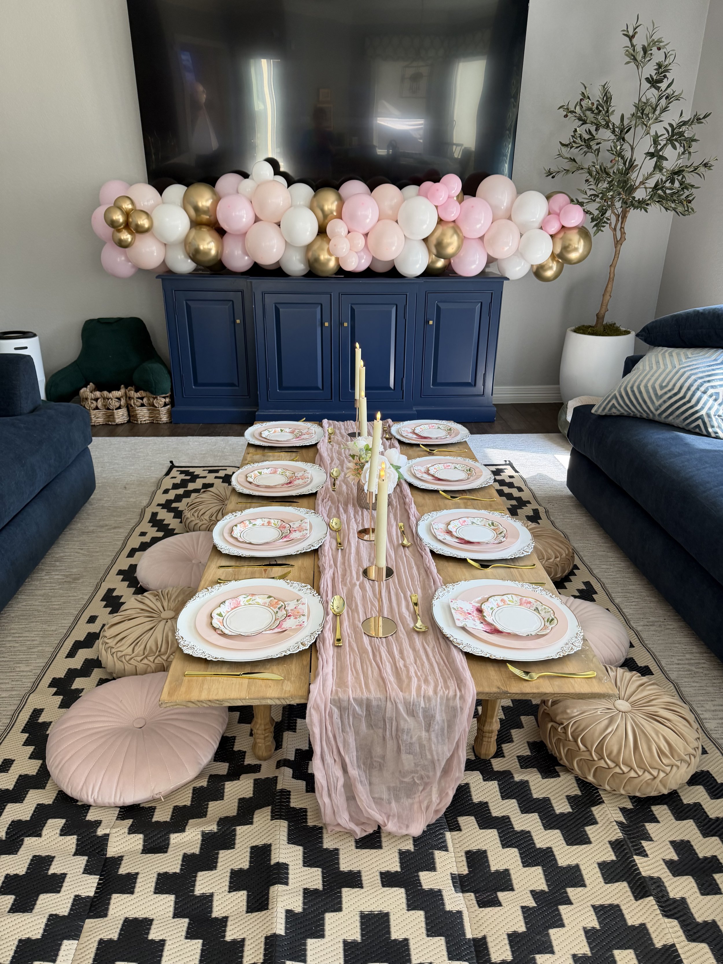 A dining table with floral dinnerware, gold and pink accents, surrounded by round pink and beige floor cushions, a garland of pink, white, and gold balloons on a blue sideboard decorated for a celebration, and a black-and-white geometric rug.