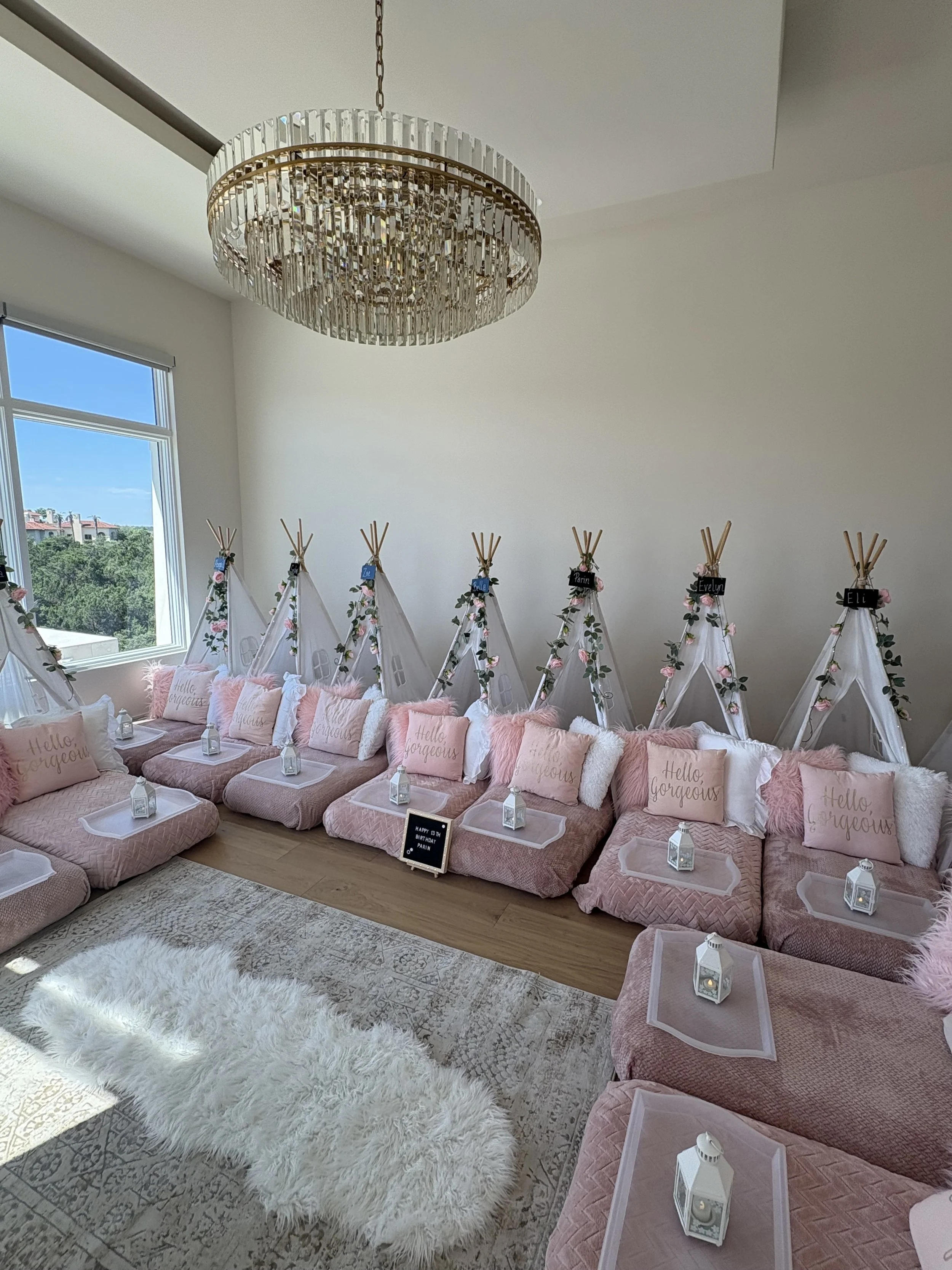 A decorated indoor space with pastel pink cushions on floor seating, teepees with floral embellishments and name tags, a white fluffy rug, a white textured sofa, a chandelier, and a large window showing a view of trees and a building.