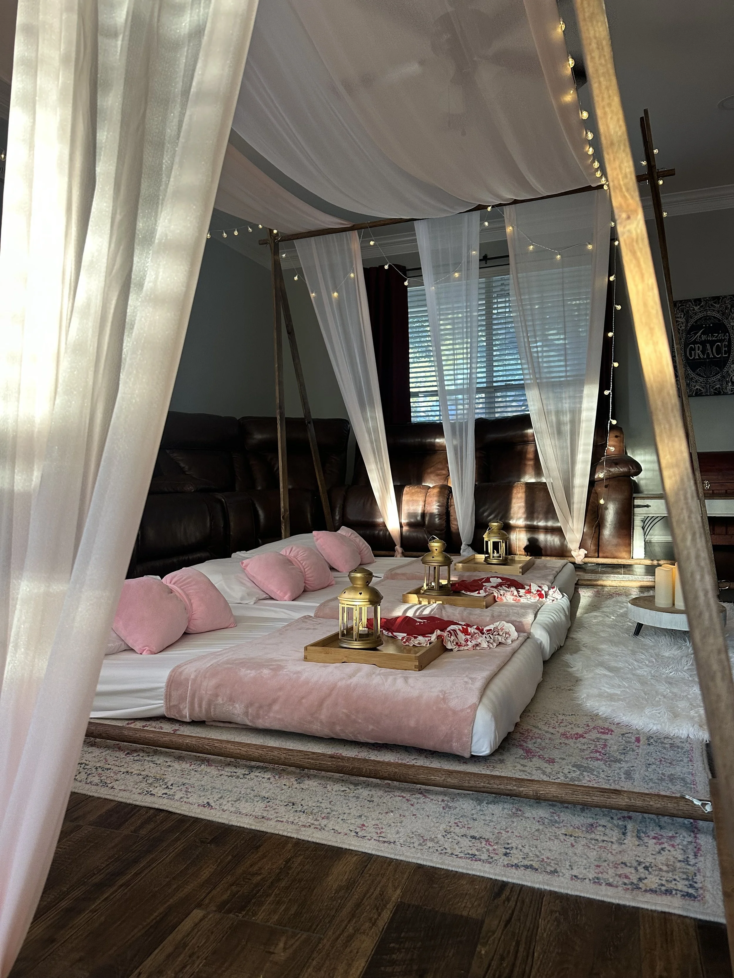 A cozy bedroom setup with a low bed decorated with pink pillows and blankets, surrounded by a wooden canopy frame draped with sheer curtains and string lights, with a patterned rug on wooden flooring and a window with blinds in the background.