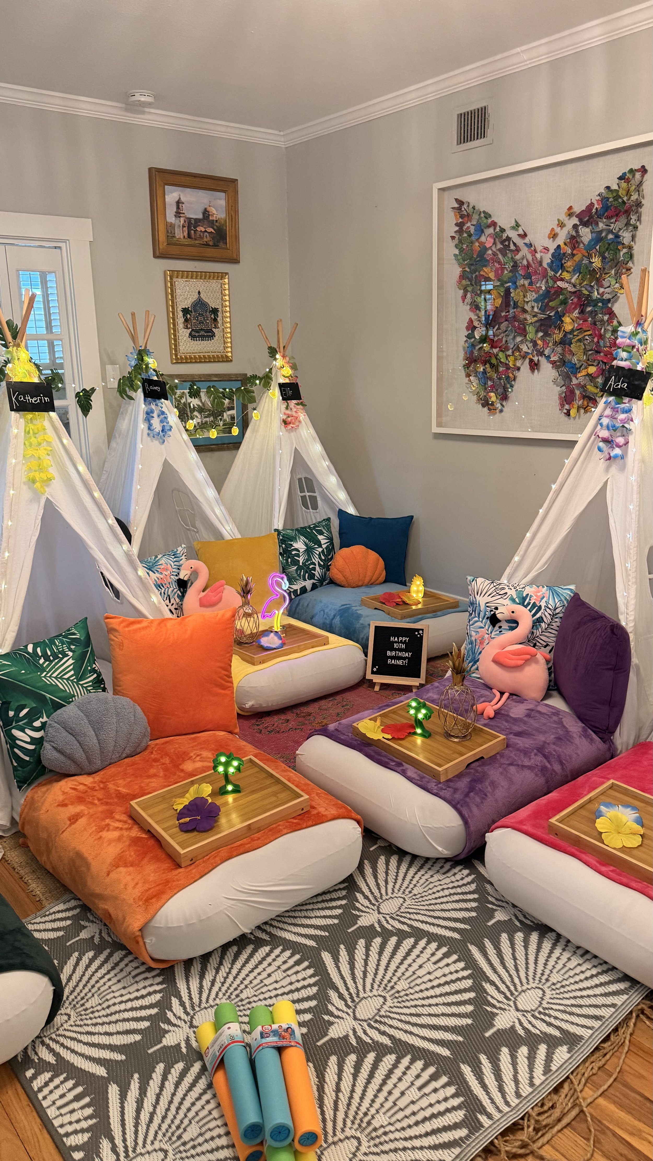 A cozy indoor space decorated for a birthday celebration with colorful pillows, stuffed flamingos, small tents, and snacks on wooden trays. There's a large butterfly artwork on the wall and soft lighting creating a festive atmosphere.