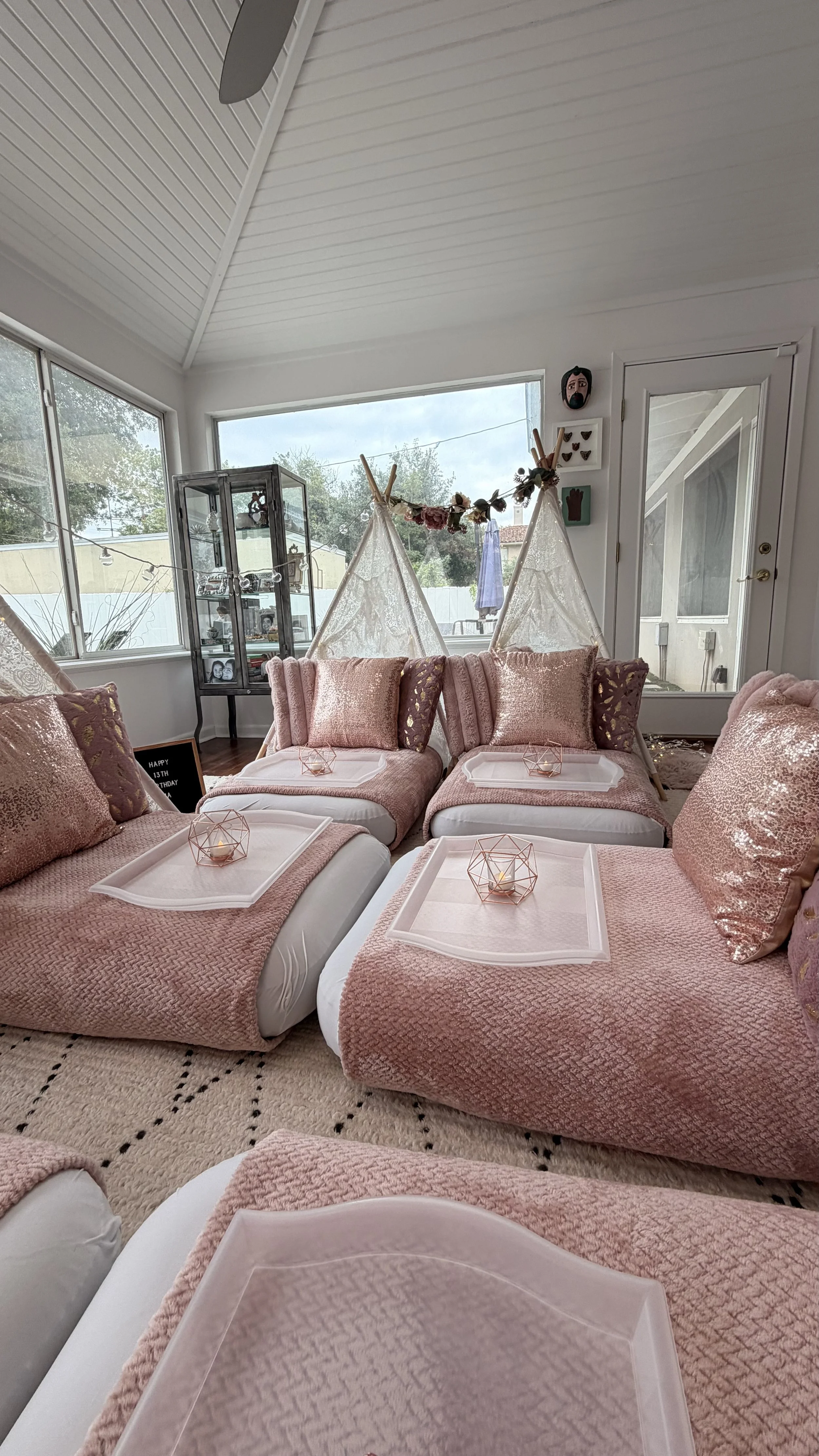 A cozy living room with pink textured blankets and sequin pillows on white beds or lounge chairs. Four pink geometric candle holders with candles are placed on each white tray, which are on top of the pink blankets. There are large windows allowing n