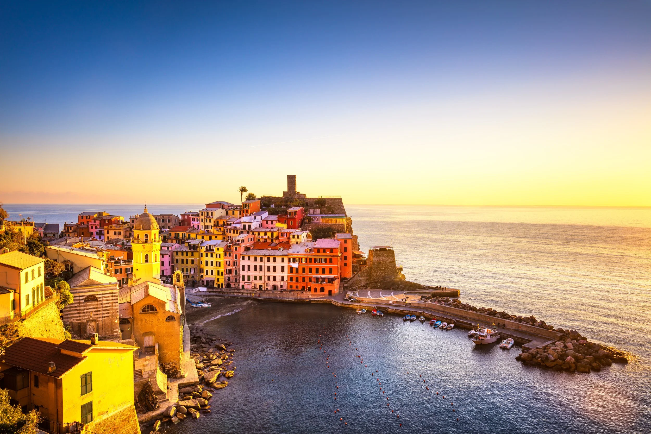 Colorful seaside village at sunset with pastel-colored buildings, a small harbor with boats, and a historic tower on a cliff.