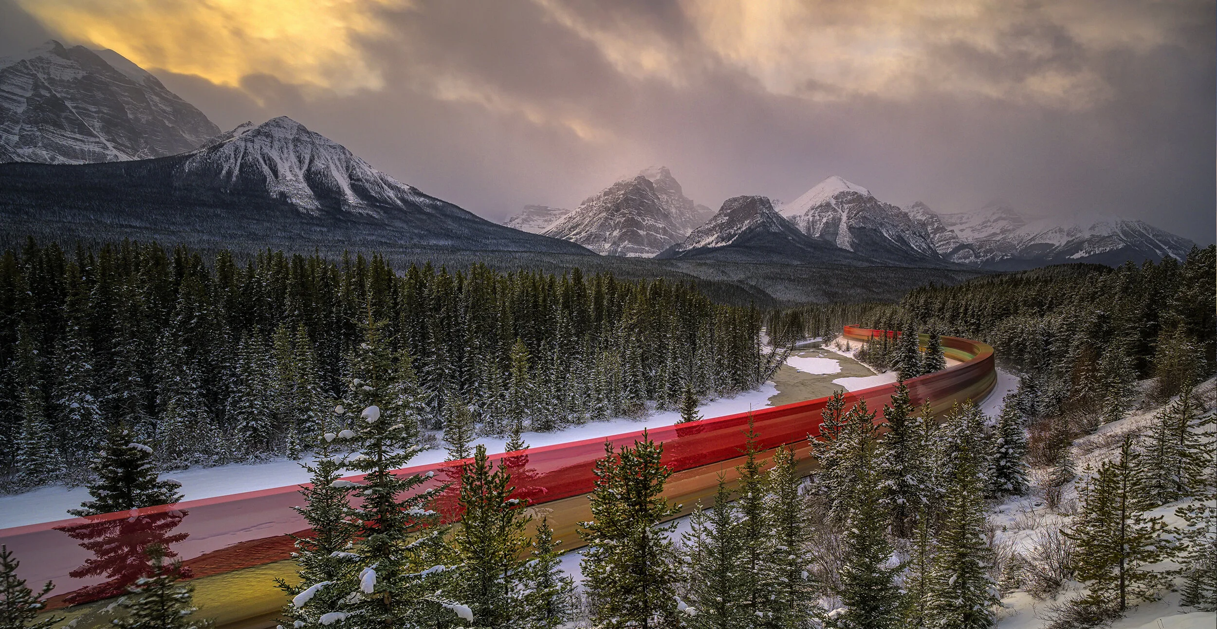 A snow-covered landscape with dense pine trees, tall mountains with snow caps in the background, and a winding railway track with red, yellow, and green blur effects through the forest during sunset.