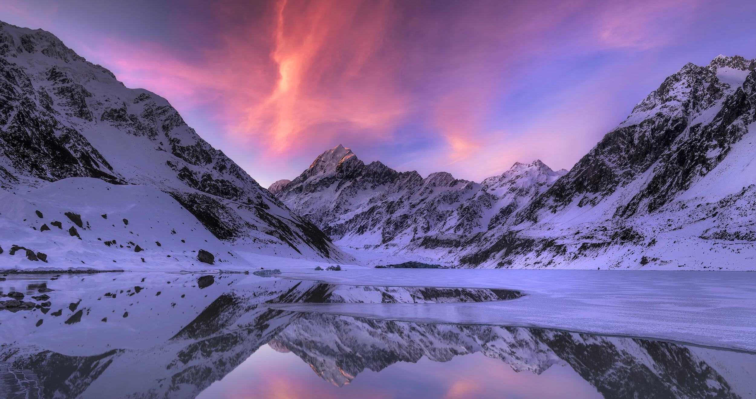 Snow-covered mountain range reflected in a calm, partially frozen lake at sunset with pink, purple, and orange sky.