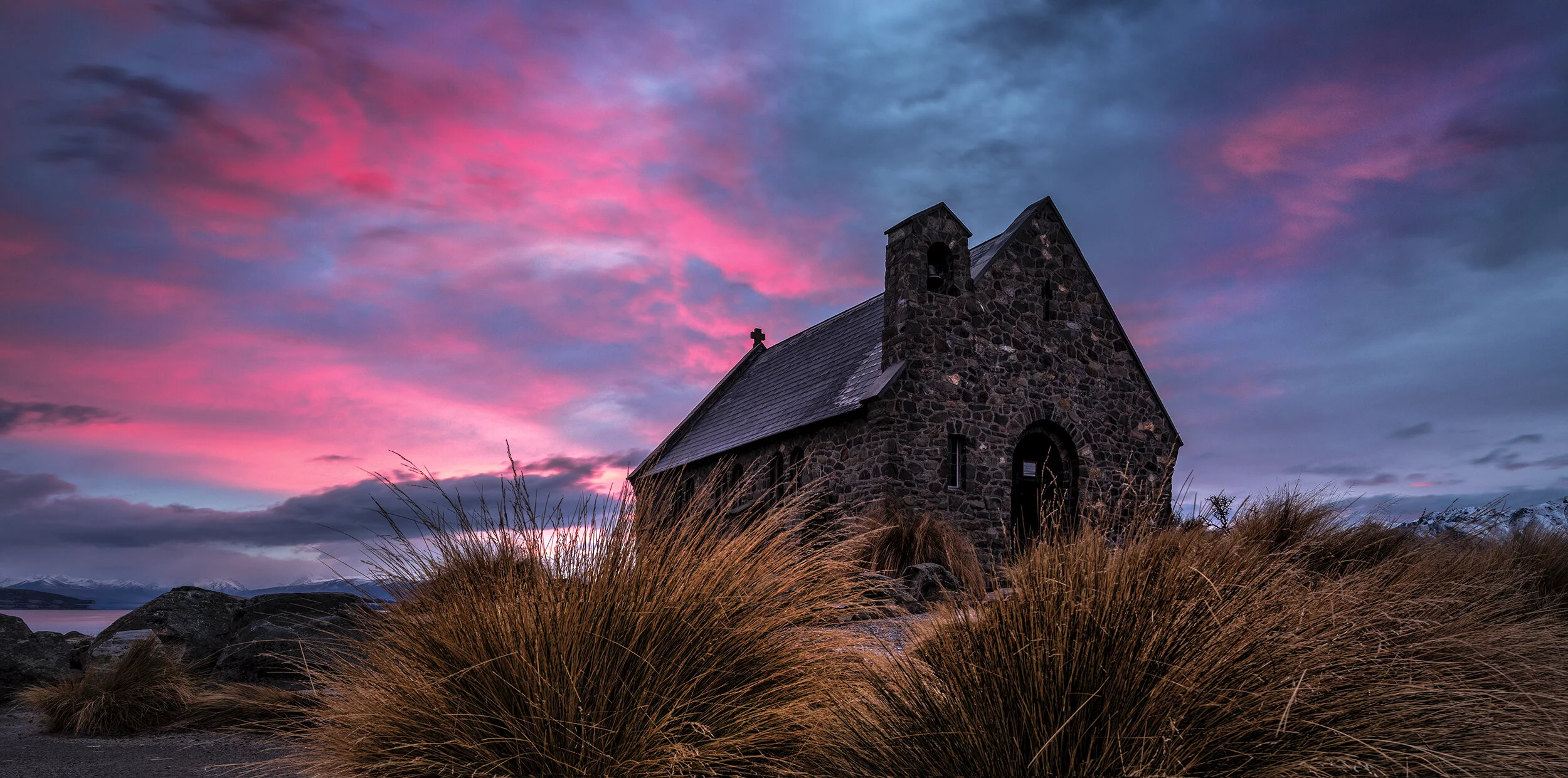 Stone church under a colorful pink and blue sunset sky with tall grass surrounding it.